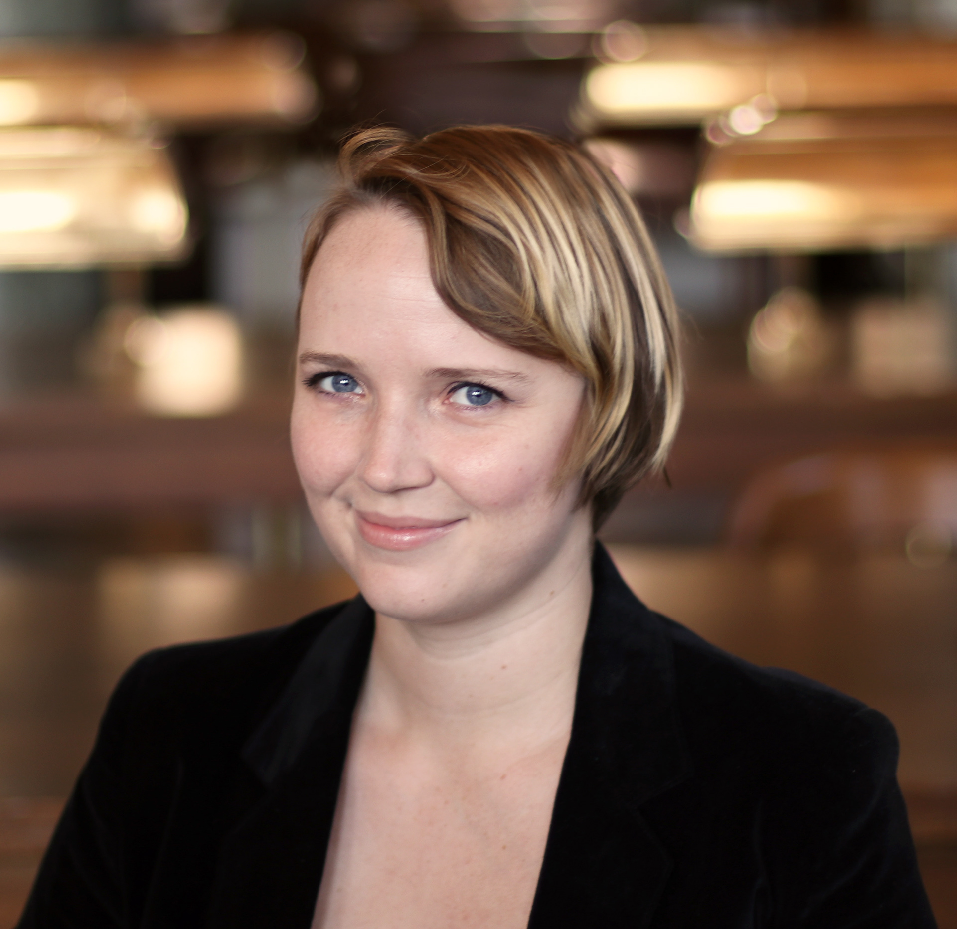 A smiling woman in her early 20s posed for a headshot. She is wearing a blazer and appears to be in a library.