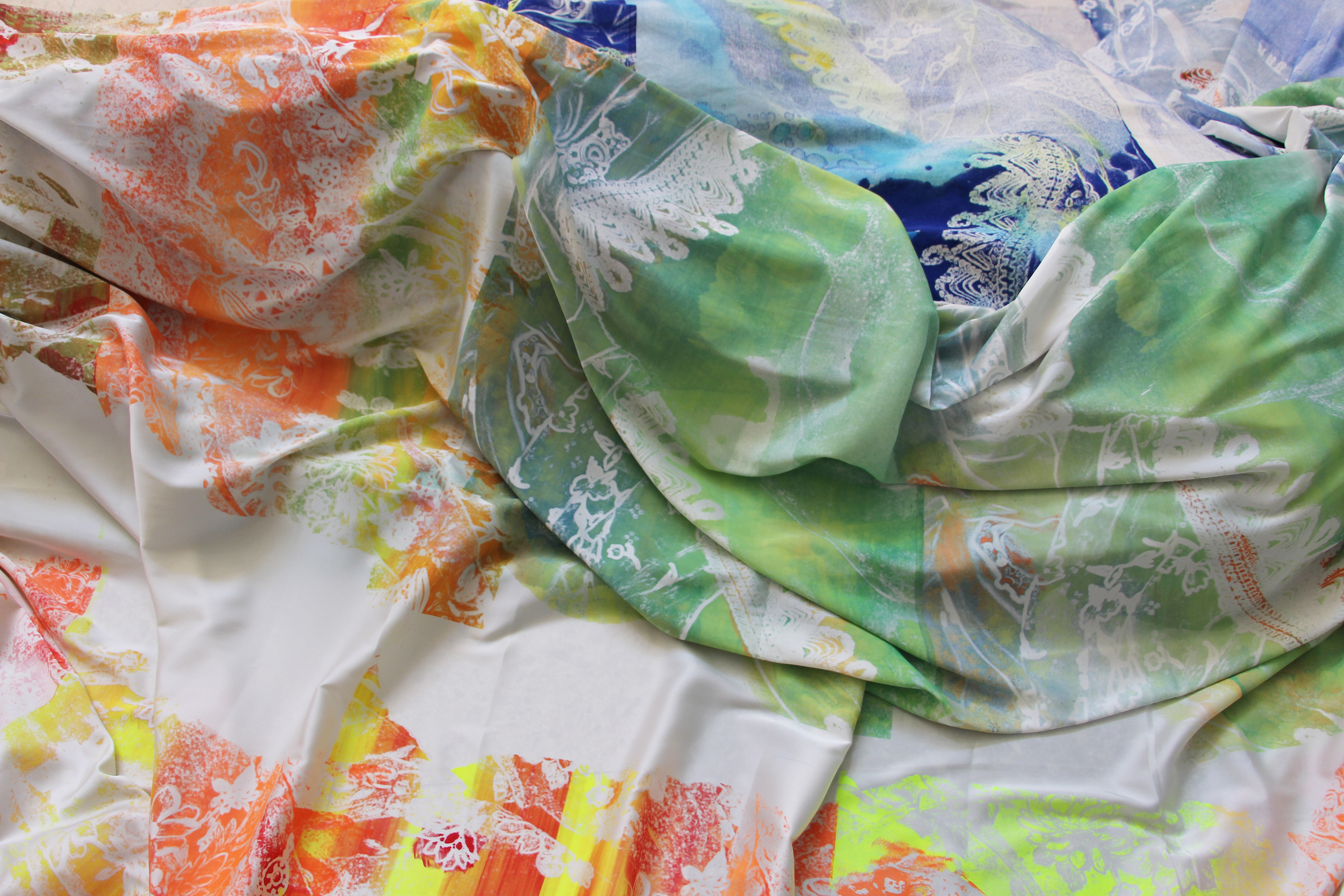 Tissu coloré aux motifs amazighs réalisés en sérigraphie et aquarelle, avec des teintes dégradées de vert, bleu, orange et jaune, et des motifs blancs délicats sur une surface souple et plissée.