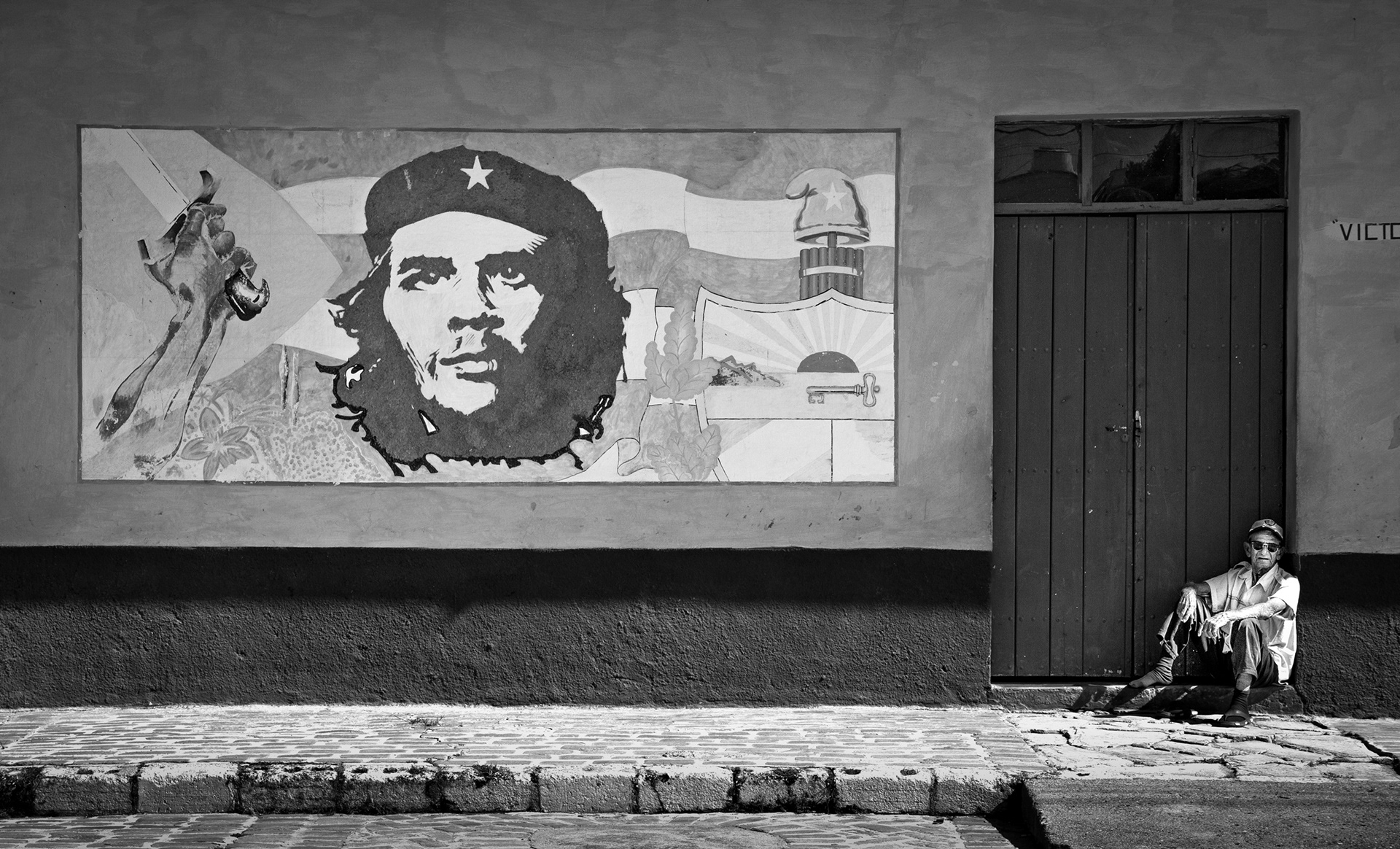 Black and white street photograph of a man sitting beside a door in Havana with a Che Guevara mural on the wall