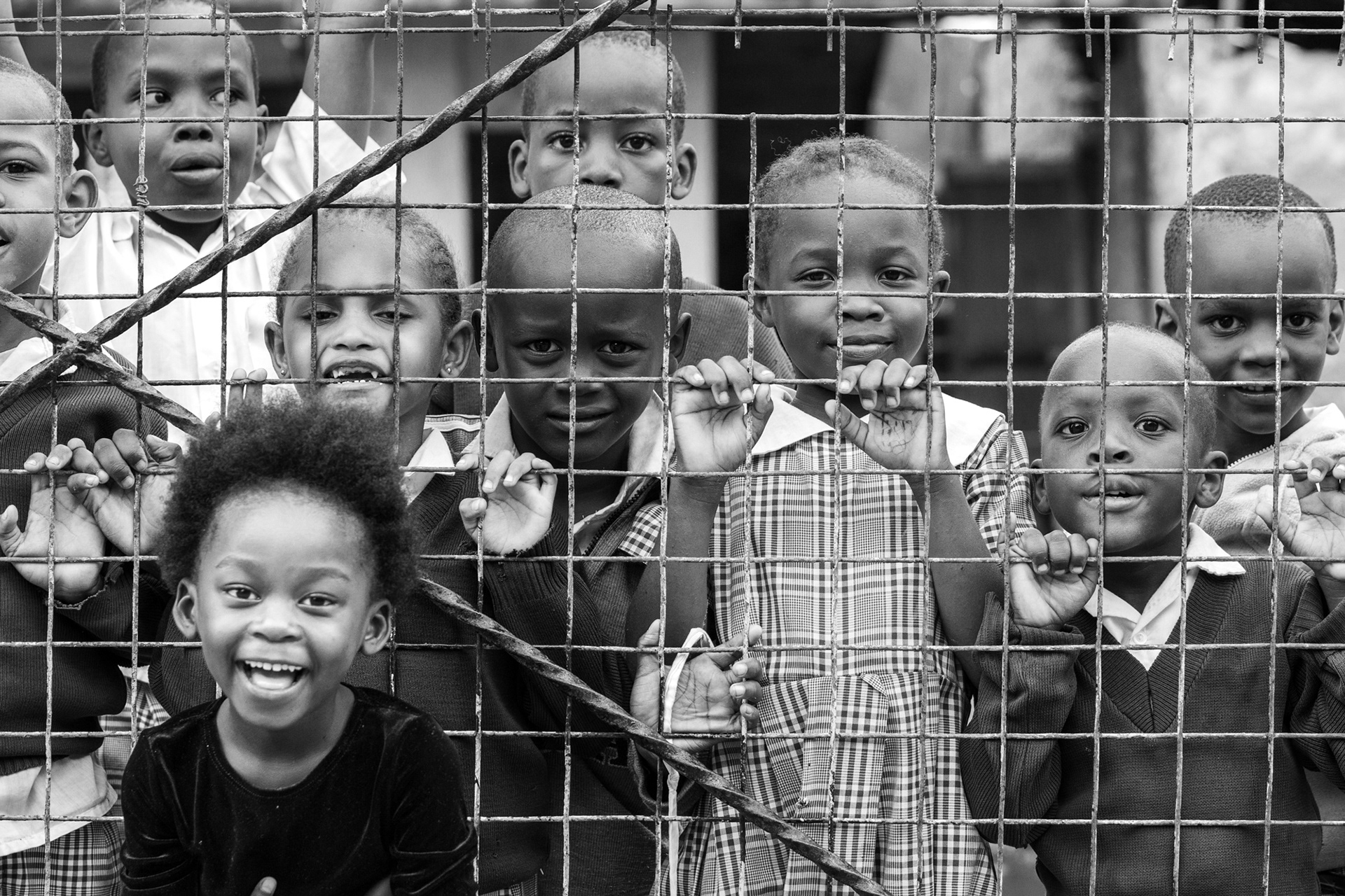 Black and white documentary photograph of a group of Kenyan school children standing behind a wire fence, looking curiously and smiling at the camera. The image captures childhood, community, and everyday life in rural Kenya, East Africa.