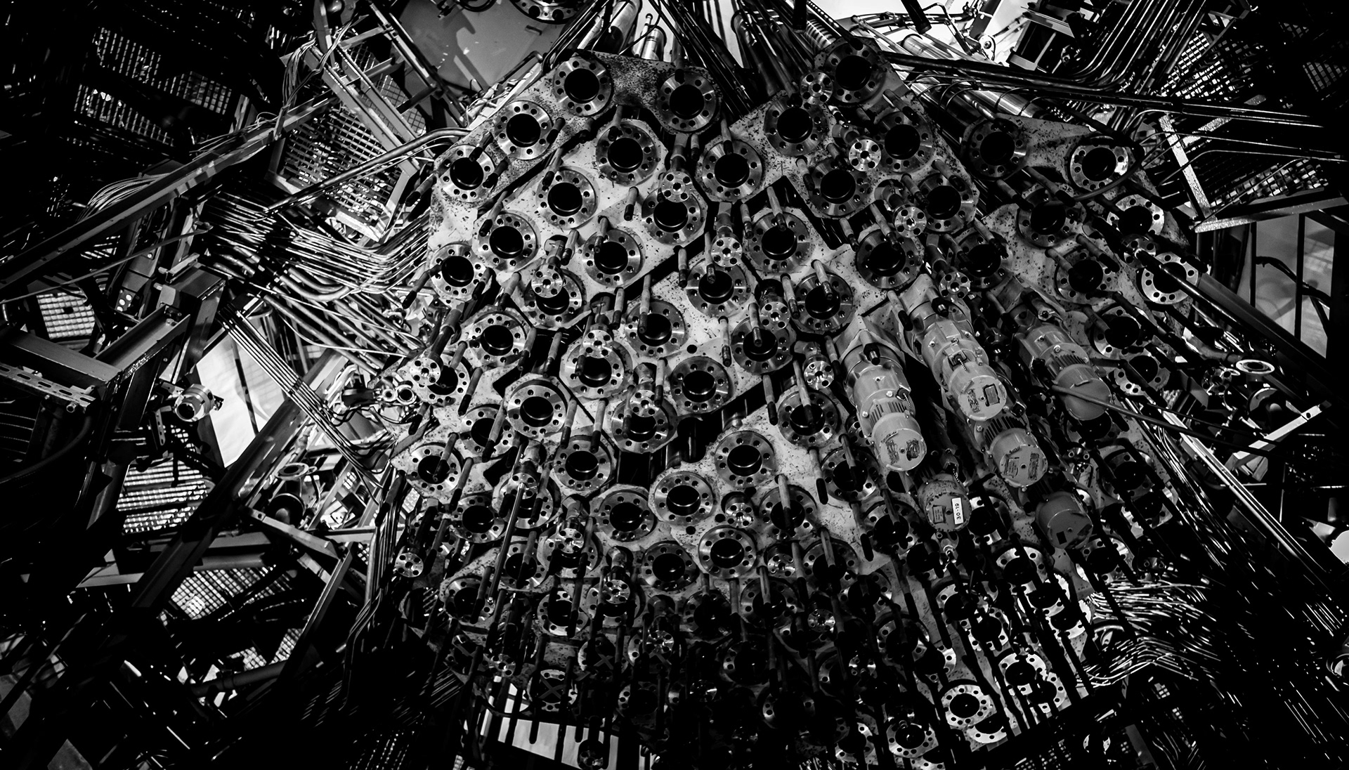 Black and white photograph of a nuclear reactor core with a dense arrangement of circular openings, pipes and cables, creating an abstract industrial pattern with strong contrast and dramatic perspective.