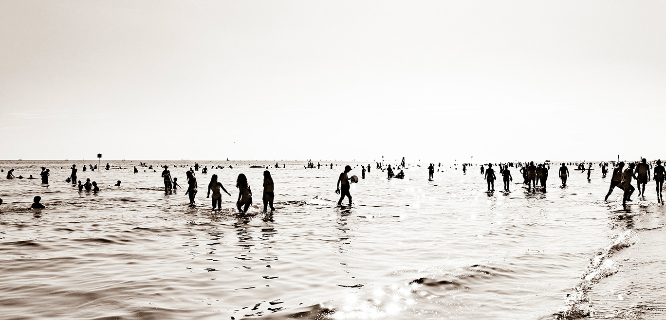 Wide beach scene in Grado with many people standing and walking in shallow water beneath a bright, open sky.