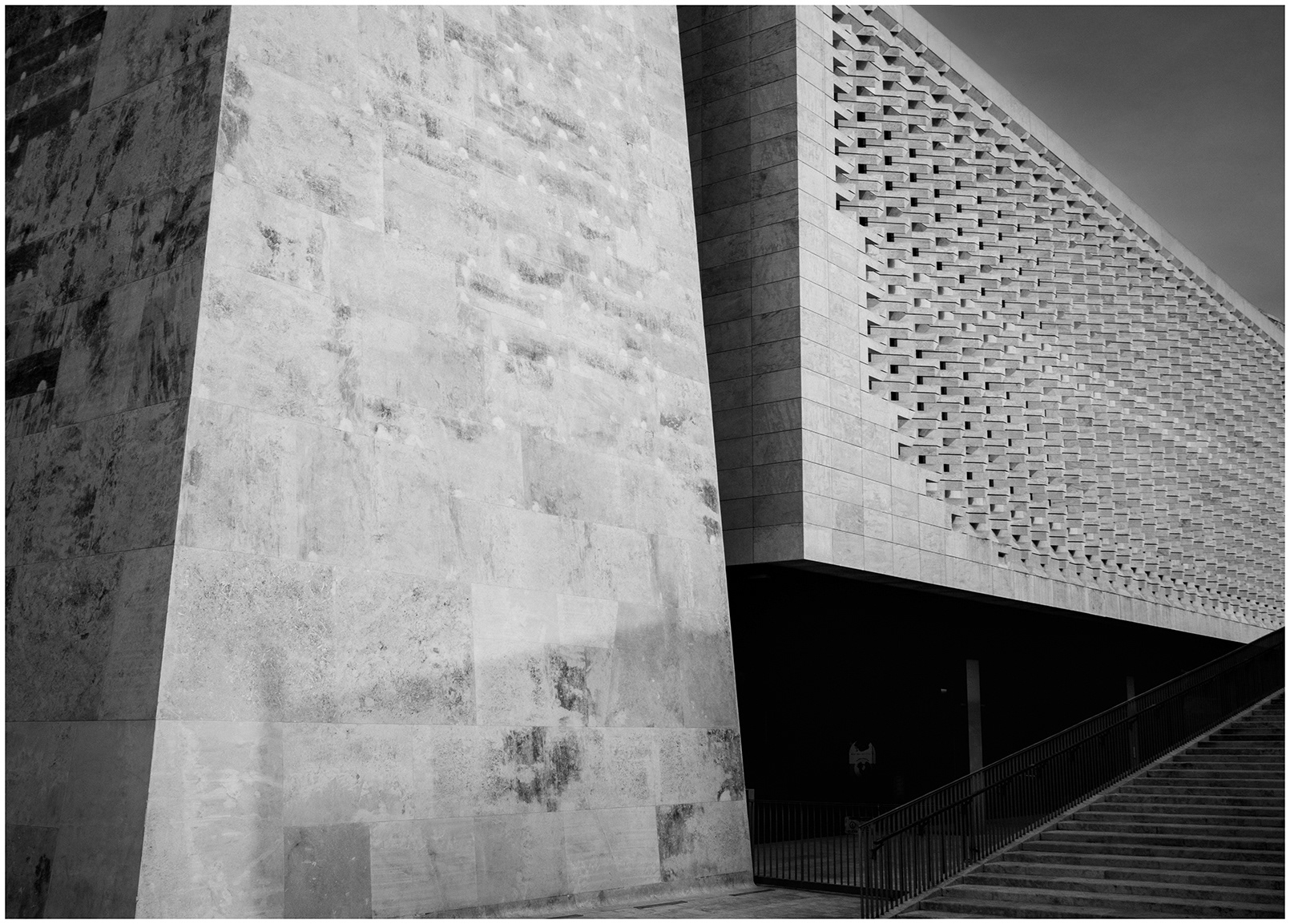 Minimalist black and white fine art architecture photograph captured in Valletta, Malta. Strong geometric lines, textured stone walls, dramatic shadows and a modern staircase create a quiet and timeless urban composition focused on structure, symmetry and silence.