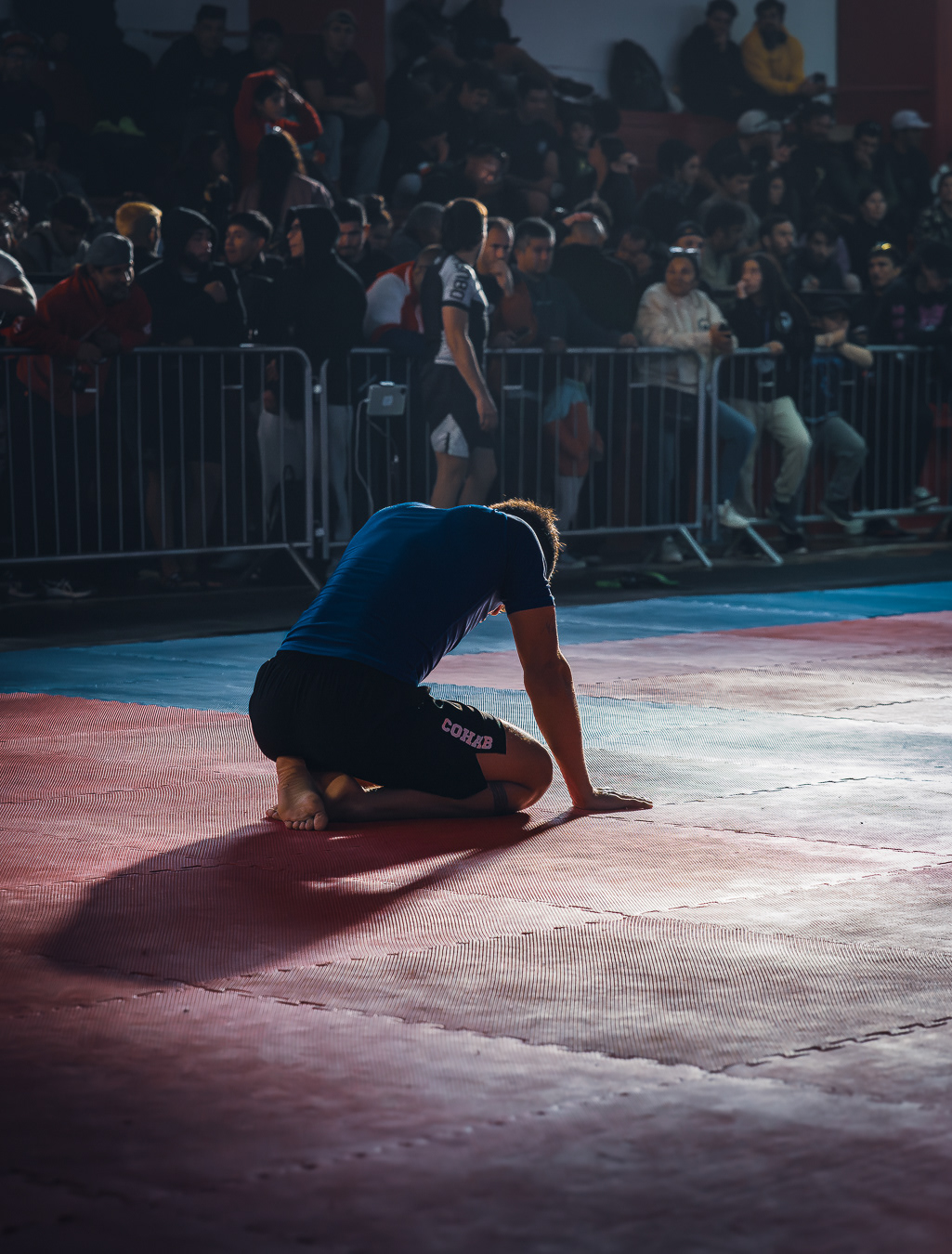 Un deportista, agotado o pensativo, arrodillado en una colchoneta de lucha en medio de un torneo, con el público en las gradas observándolo.