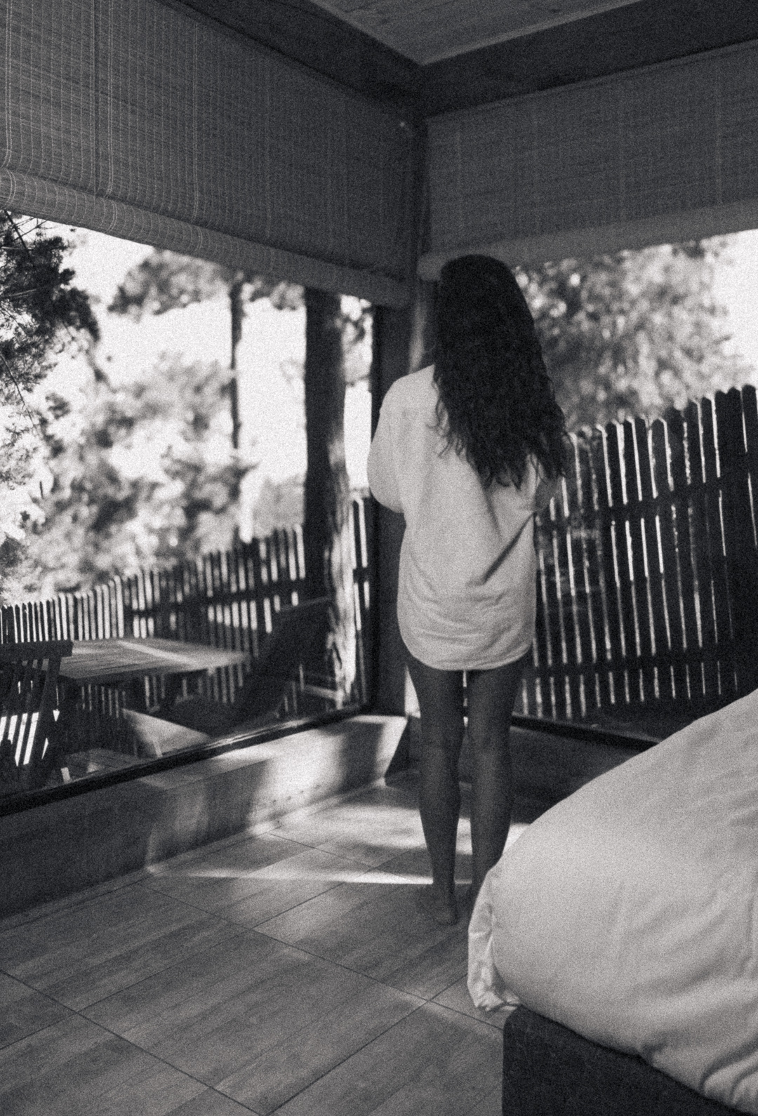 Una mujer vista desde atrás, de pie en un porche o habitación luminosa, mirando a través de una ventana al bosque. Está descalza y lleva una camisa blanca y larga.