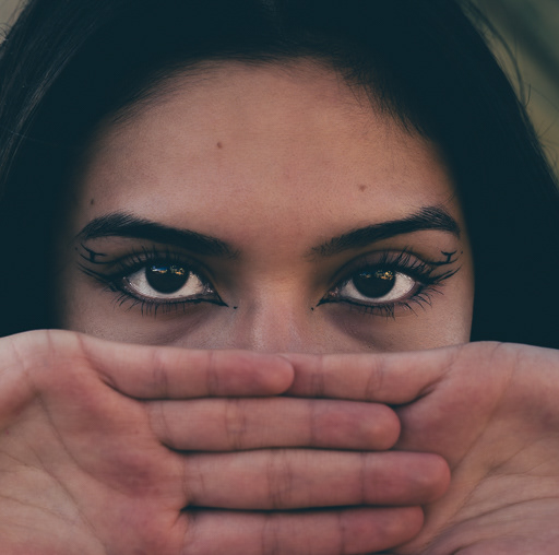 Una toma en primer plano del rostro de una mujer. Sus ojos están abiertos y mirando directamente al frente, con un delineado artístico que se extiende más allá de sus pestañas. Sus manos están entrelazadas cubriendo la mitad inferior de su cara, desde la nariz hasta la barbilla