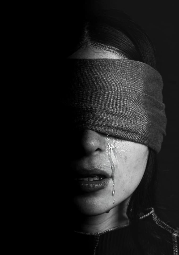 Una foto en blanco y negro de una persona con los ojos vendados, con una lágrima bajando por su mejilla. La imagen está capturada con una iluminación dramática, con la mitad izquierda del rostro en sombra total
