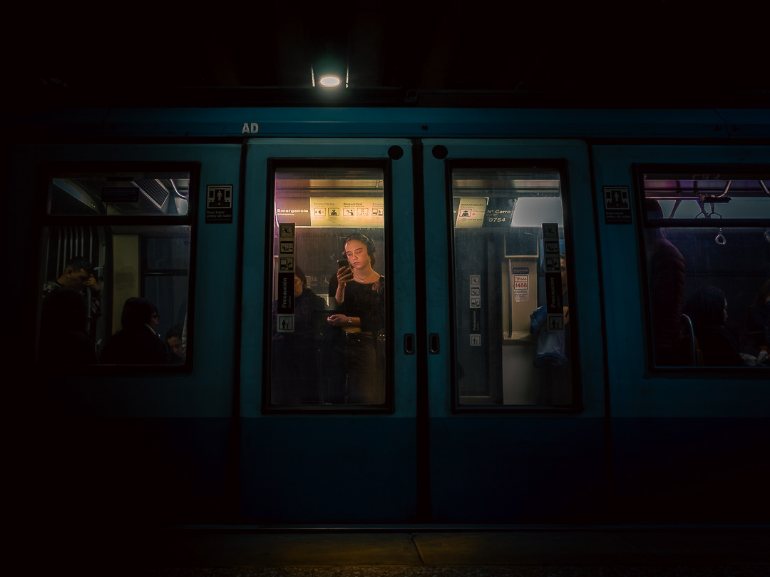 Escena nocturna en un andén de metro donde se ve el exterior de un vagón azul. A través de la ventana central, una mujer con auriculares observa su celular, destacándose por la luz interior frente a la penumbra del entorno