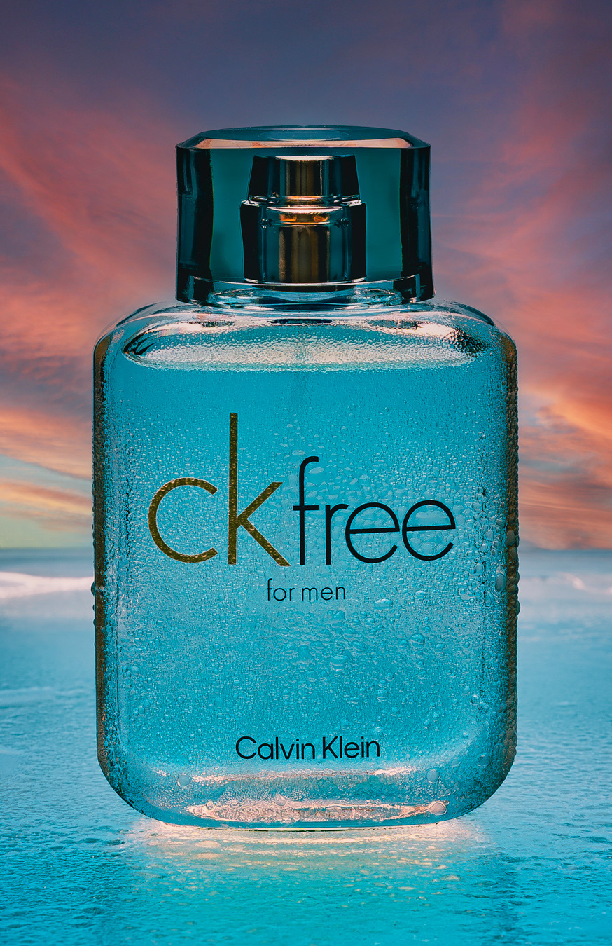  Un frasco de perfume "CK Free for Men" de Calvin Klein, cubierto de gotas de agua, con un fondo de atardecer.