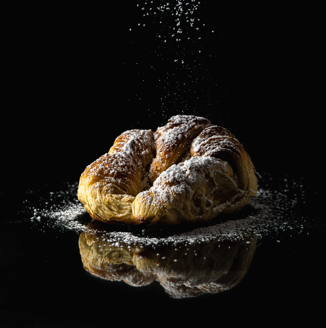 Una factura, posiblemente un croissant o medialuna, espolvoreada con azúcar en polvo, está fotografiada sobre una superficie brillante que refleja su imagen. El fondo es completamente negro, lo que hace que la iluminación se centre en el producto. El azúcar en polvo parece estar cayendo sobre la factura, creando un efecto de movimiento