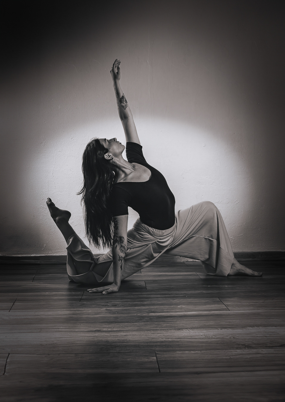 Una mujer vestida de negro y pantalones anchos realiza una pose de yoga. Su rodilla izquierda está en el suelo mientras estira la pierna derecha hacia atrás, y tiene su brazo derecho extendido hacia arriba. La foto está en blanco y negro con tonos azules.