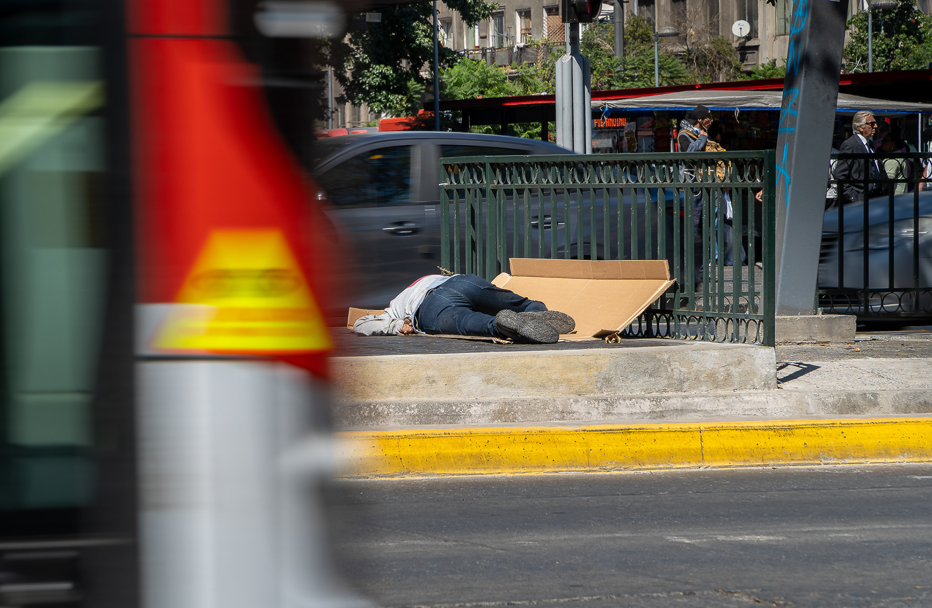 Fotografía urbana con enfoque en una persona en situación de calle durmiendo de lado sobre cartones junto a una reja metálica en la vereda. En primer plano desenfocado pasa un autobús rojo y amarillo, mientras de fondo se aprecia el tráfico vehicular y transeúntes.