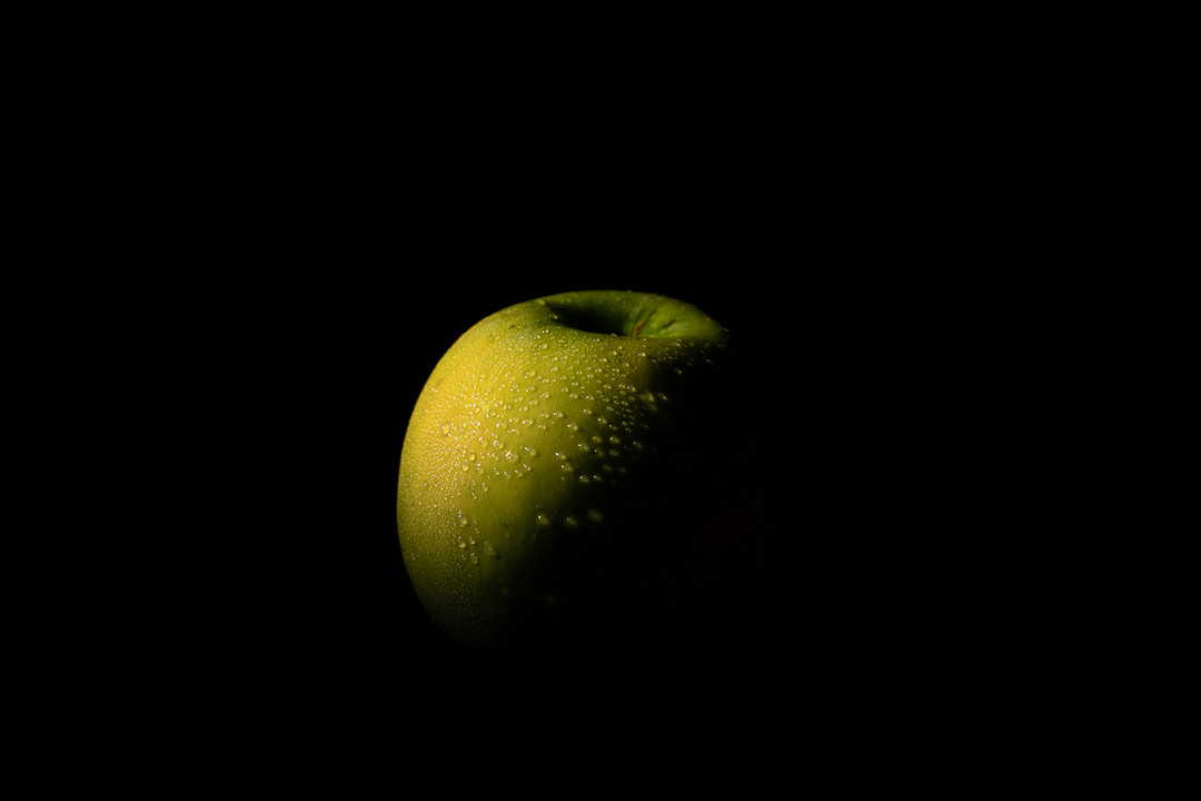 "Fotografía publicitaria de manzana verde con gotas de agua sobre fondo negro, técnica de iluminación claroscuro."