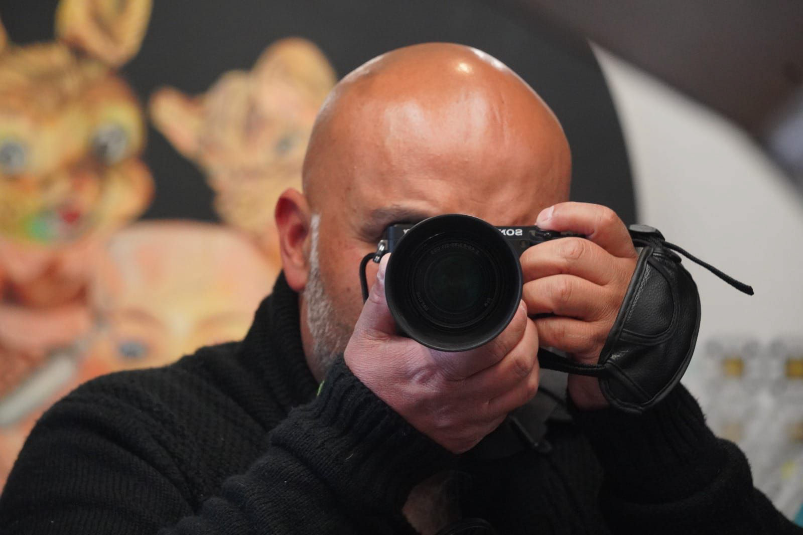 Una toma en primer plano de Rafael Macklins, fotógrafo de Busca tu Foto, sosteniendo una cámara negra Sony y apuntando directamente al espectador. El hombre calvo con barba gris está vistiendo un suéter de punto negro. El fondo está desenfocado, con figuras de animales de juguete borrosas en la parte superior izquierda