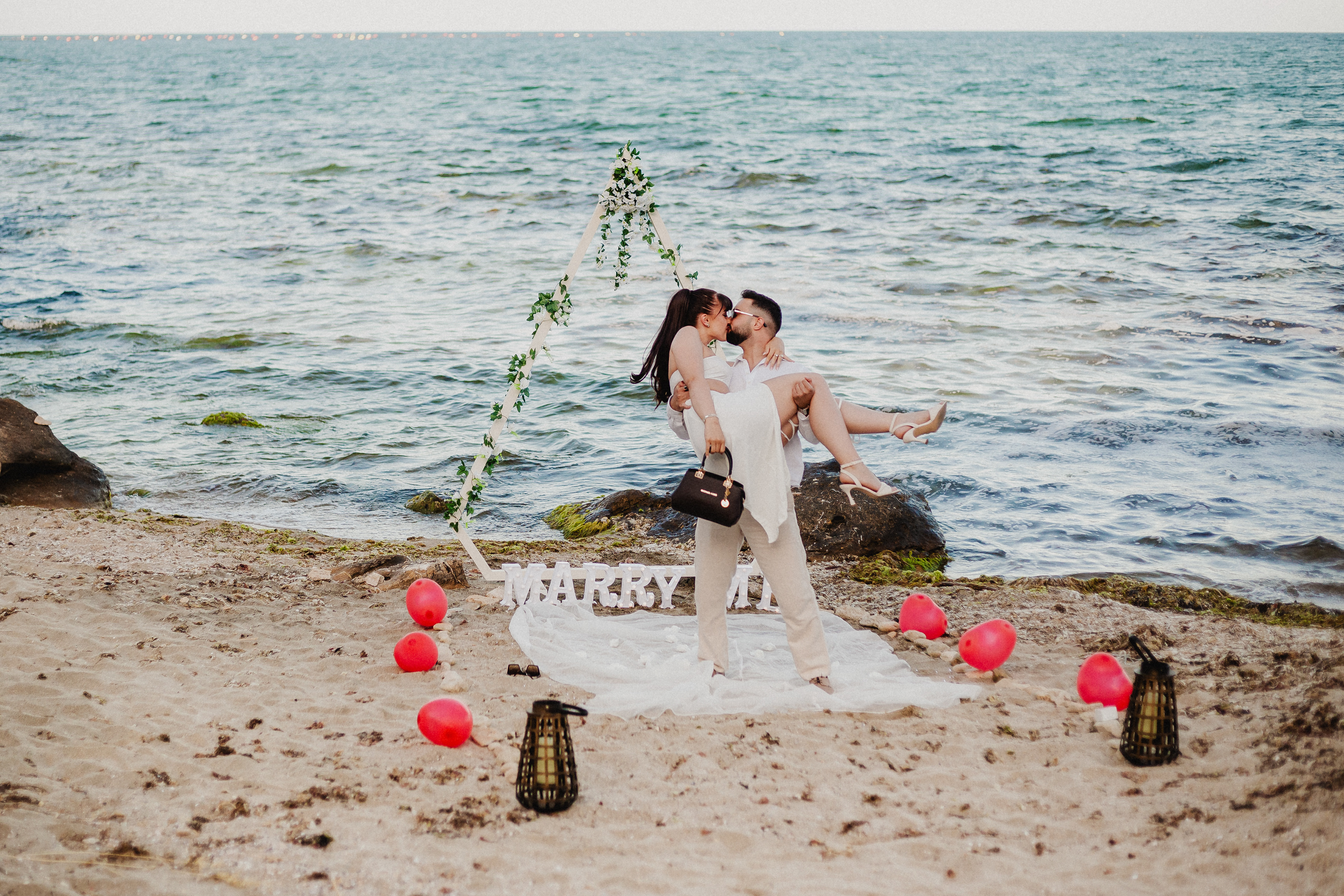 Couple celebrating a seaside proposal under a diamond-shaped wedding arch with greenery decor – romantic ‘Marry Me’ setup on a European beach at sunset.