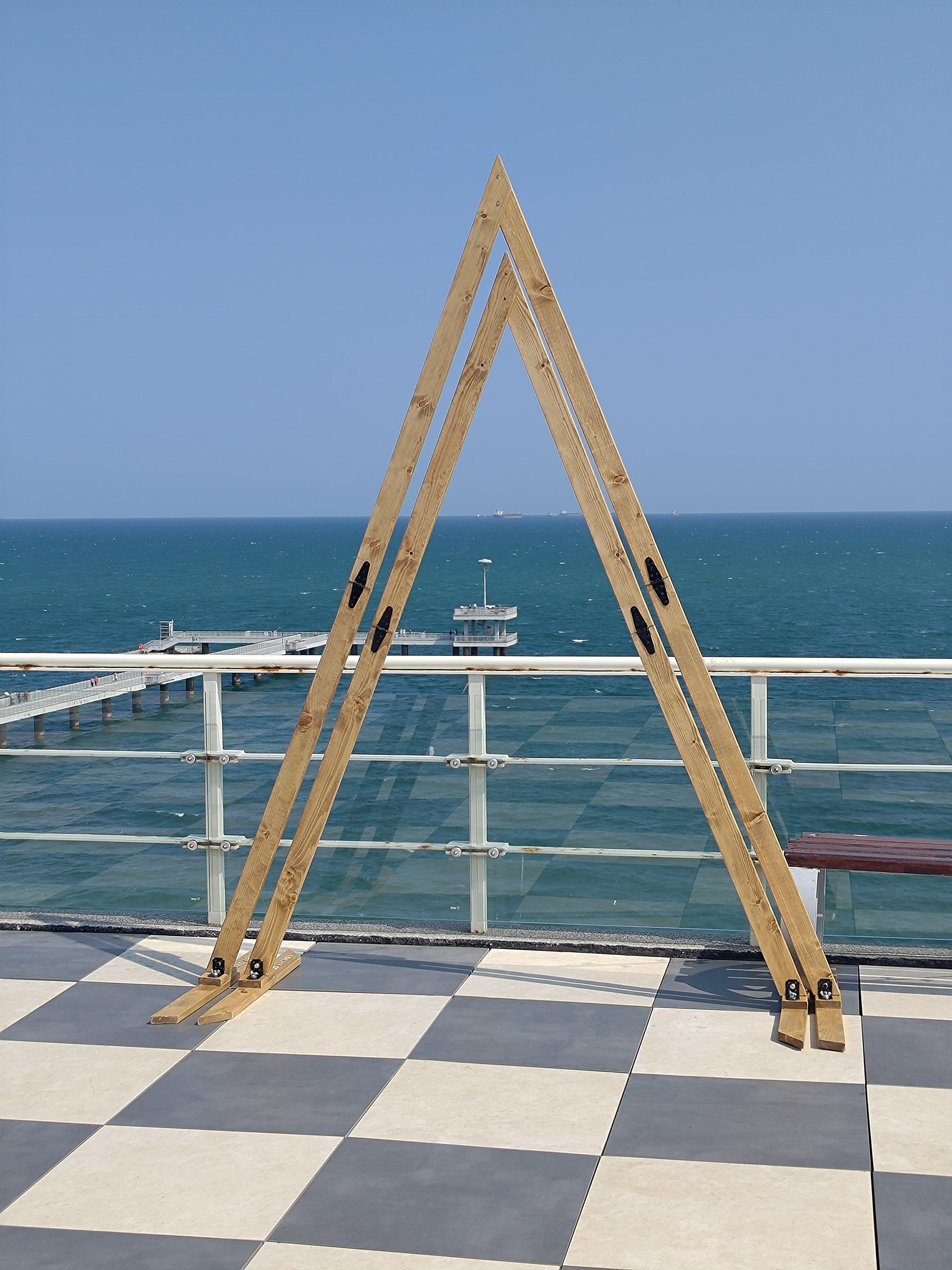 Minimalist double triangle wooden wedding arch on a rooftop with sea view – modern boho ceremony backdrop for European elopements and coastal weddings.