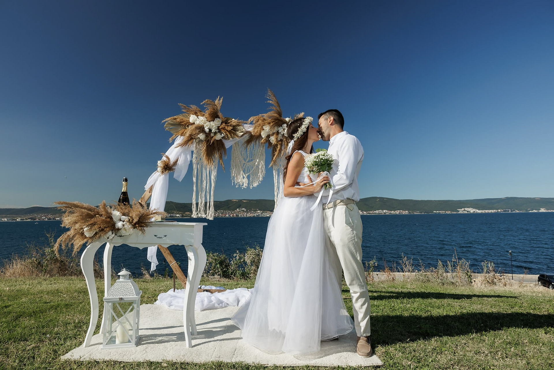 Bride and groom kissing under a boho hexagonal wedding arch adorned with pampas grass, macrame, and white drapery, set on a seaside cliff – ideal for romantic outdoor weddings and elopements in Europe.