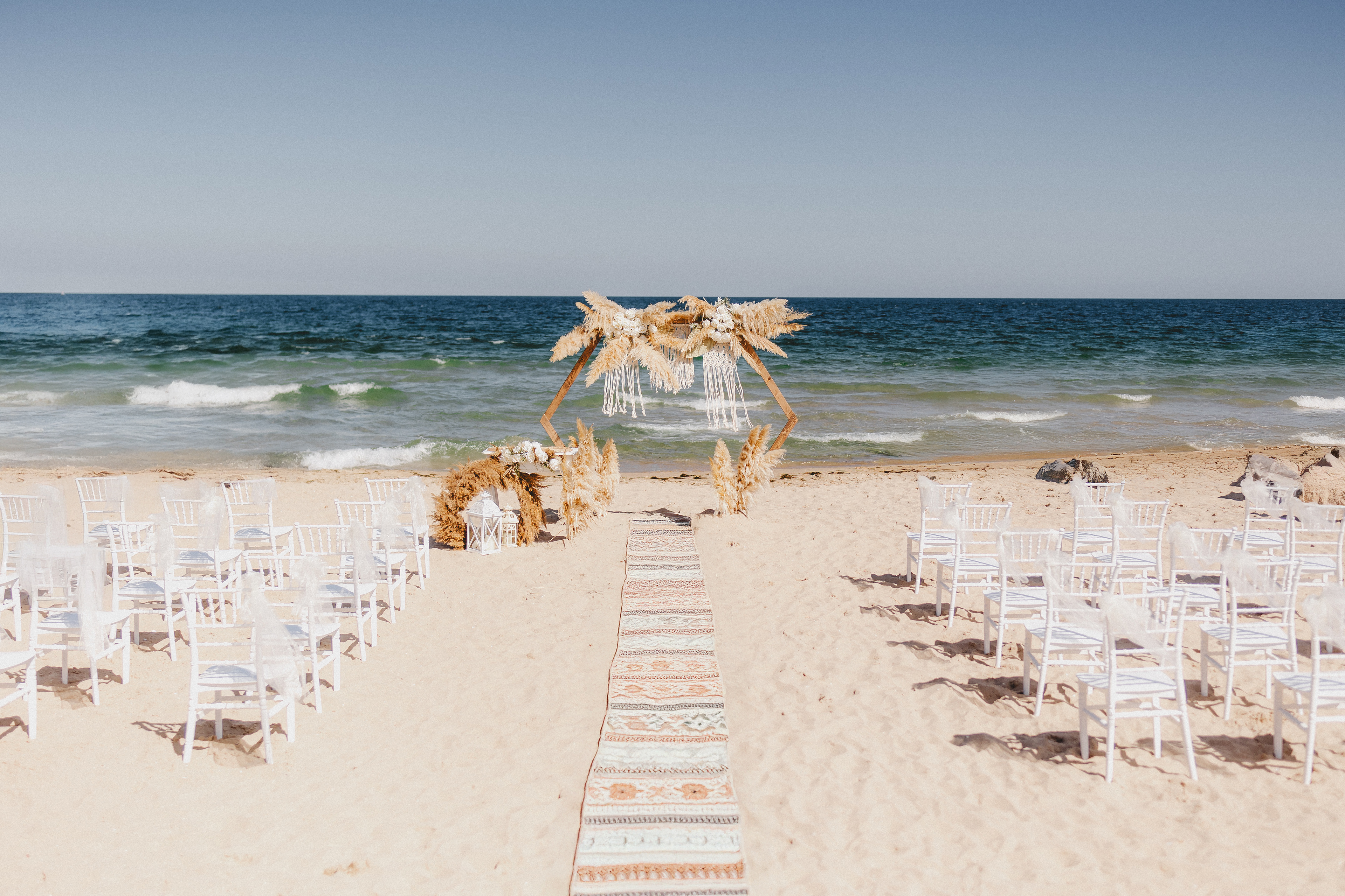 Beach wedding setup with a hexagonal wooden arch decorated with pampas grass and macrame, surrounded by white chairs and a boho-style aisle runner – perfect for seaside wedding ceremonies and coastal elopements in Europe.