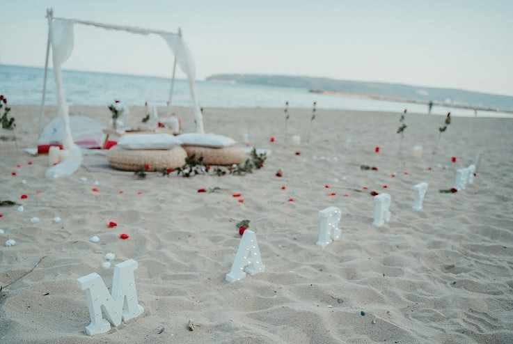 Beach proposal setup at golden hour with cozy straw pillows, floral path, and "Marry Me" letters glowing in the sand – the perfect seaside moment for a heartfelt engagement.