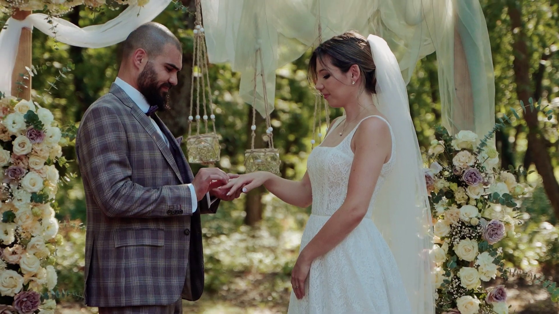 Bride and groom exchanging rings beneath a square wooden wedding arch in a forest, surrounded by floral arrangements and glass lanterns – a deeply emotional ceremony scene in nature, perfect for boho weddings in Europe.