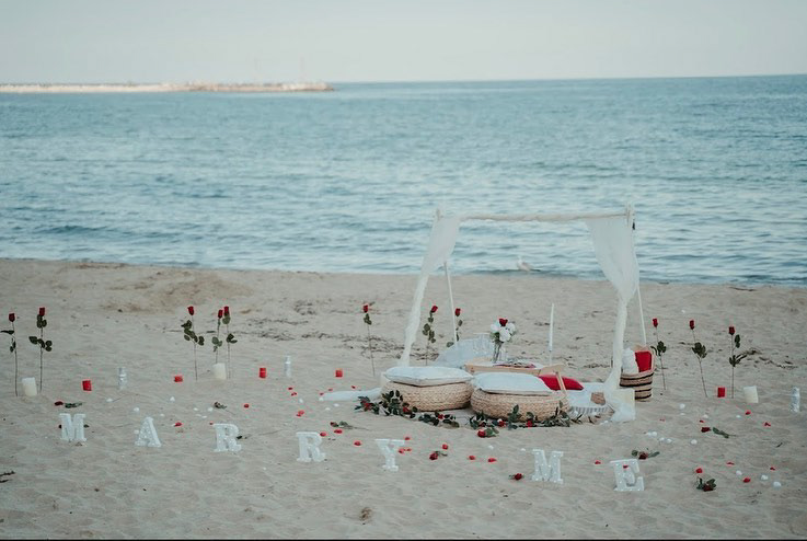 Romantic beachfront setup for a proposal at sunset, featuring a white canopy tent, rose petals, “Marry Me” letters, and cozy floor cushions facing the sea.