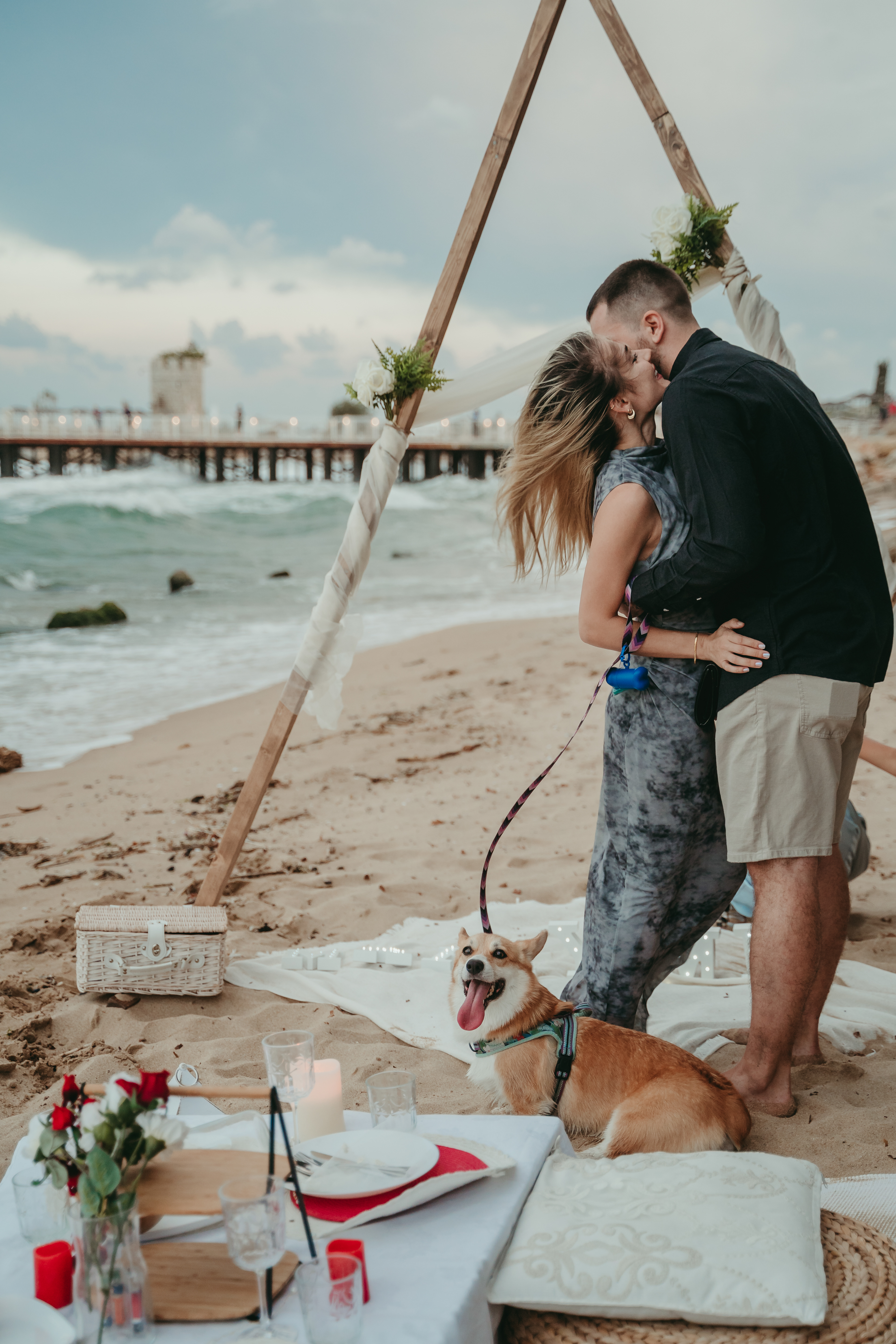 Romantic seaside proposal under a wooden triangle wedding arch, decorated with white flowers and soft fabric – a dreamy engagement moment in Europe with a happy couple and their corgi.