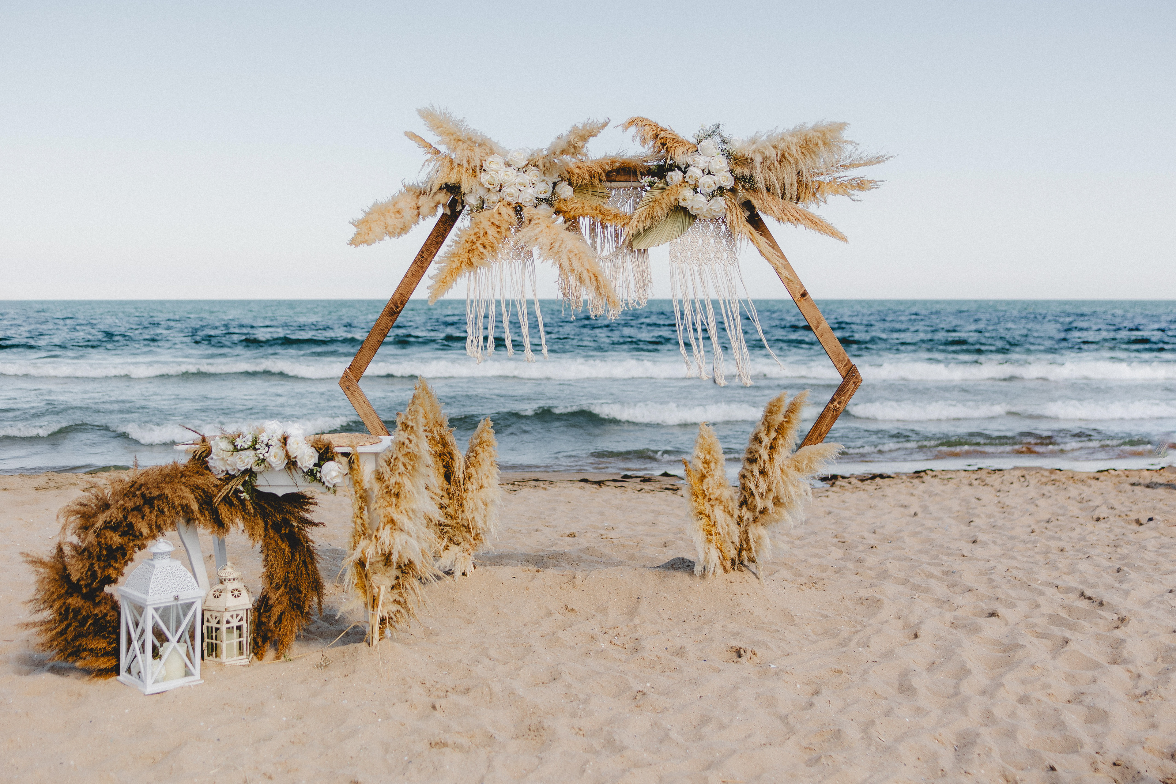 Boho hexagonal wooden wedding arch decorated with pampas grass and white roses, set on a sandy beach by the sea – ideal for coastal wedding ceremonies and destination elopements in Europe.