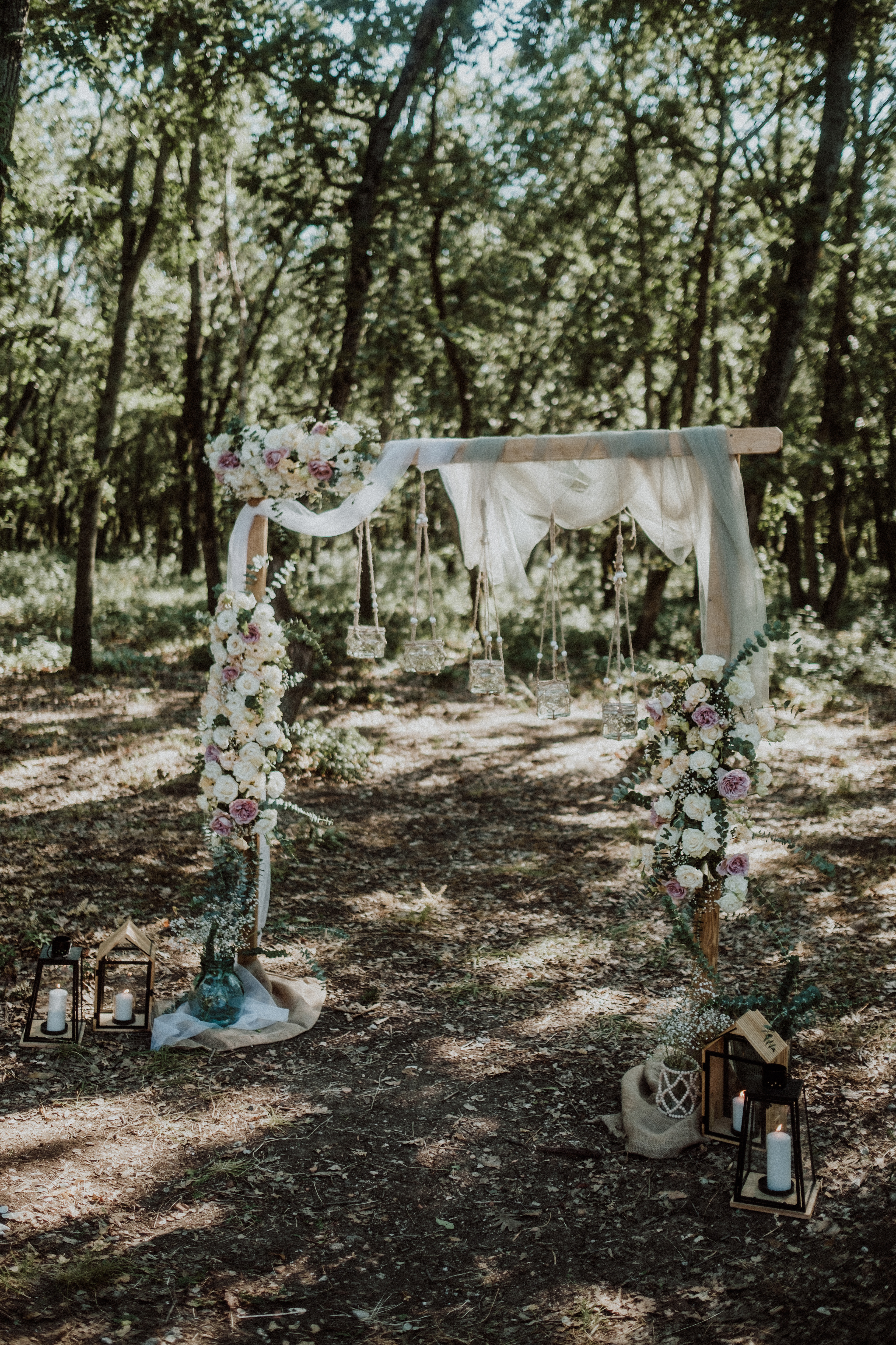 Square wooden wedding arch in a forest ceremony setup, decorated with cascading flowers, soft fabric drapes, and hanging candle jars – a whimsical boho scene perfect for woodland weddings in Europe.