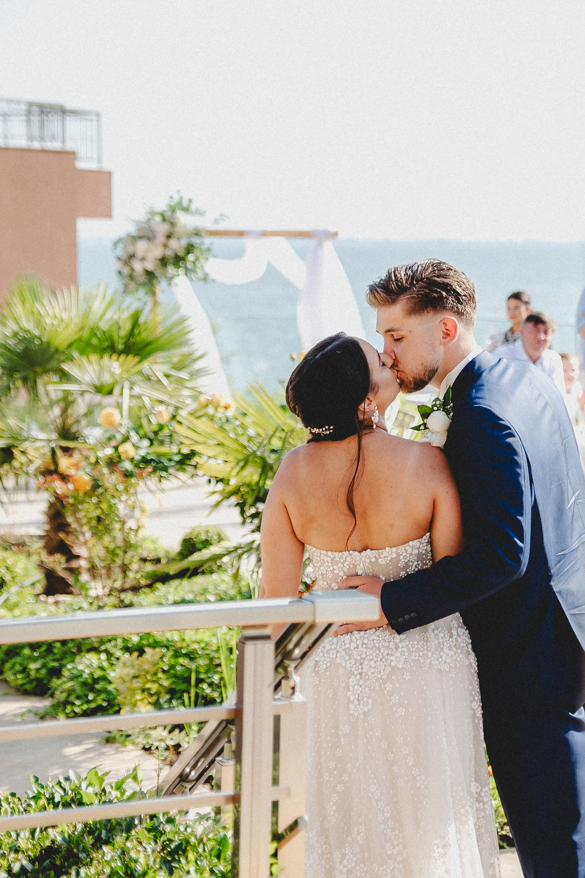 Romantic seaside wedding moment – a couple kisses after saying “I do,” framed by a square wooden arch adorned with white drapery and florals, with the sea and palm trees in the background.