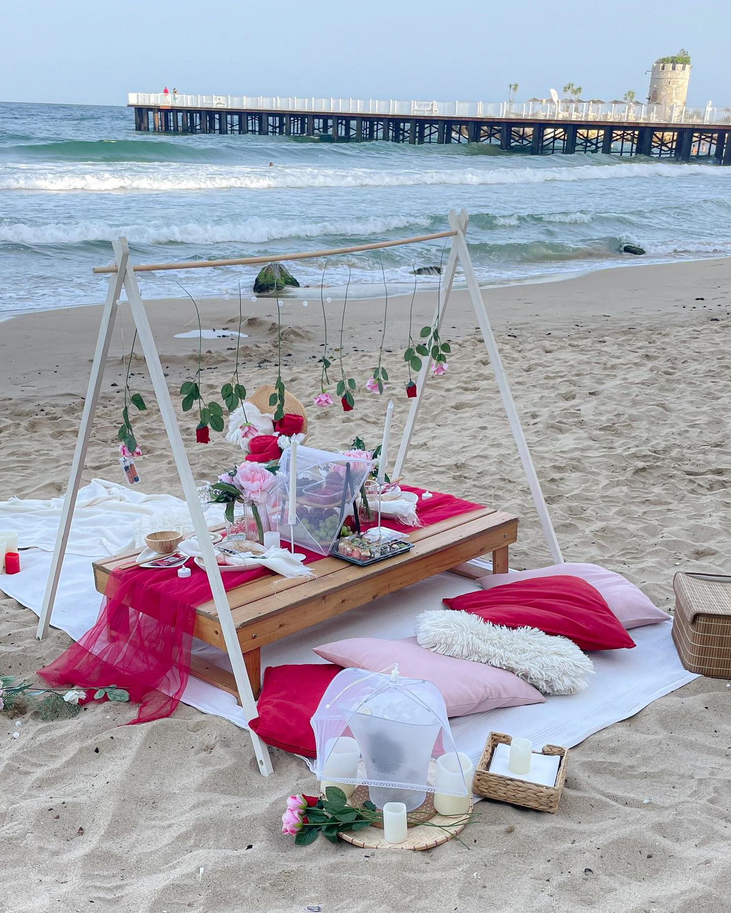 Romantic beach picnic tent setup with pink and red cushions, a low wooden table decorated with flowers and candles, and hanging roses – a dreamy scene for proposals or special celebrations in Europe.