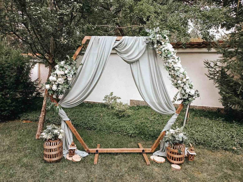 Elegant hexagonal wooden wedding arch decorated with white flowers and flowing drapery, set in a garden with rustic lanterns – perfect for romantic backyard weddings and boho ceremonies in Europe.