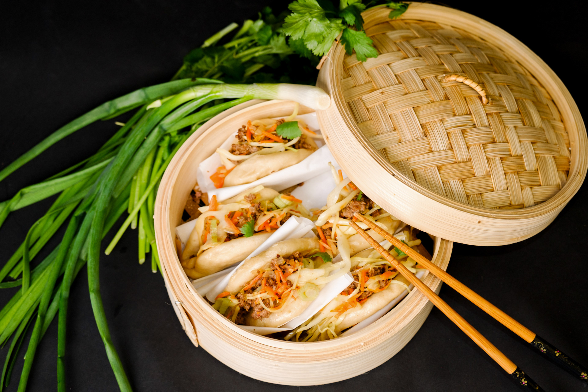 Asiatisches Essen in Bambusschüssel mit angelehntem Bambusdeckel auf dunkler Tischplatte. Produktfotografie von Georgi Sarkezi