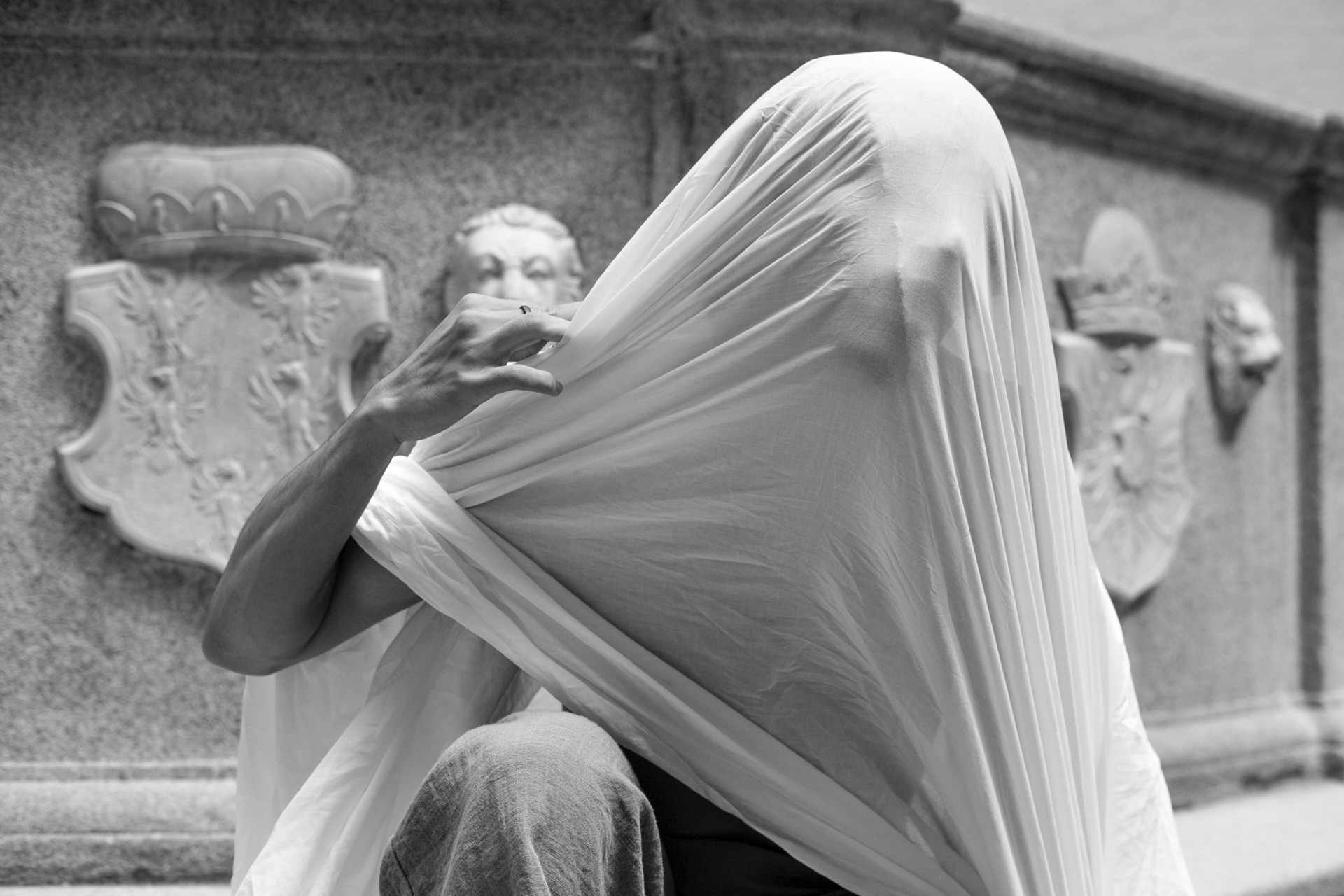 Ein Mensch am Boden vor einem Brunnen mit einem großen weißen Tuch, das seinen Kopf und Oberkörper verdeckt. Die rechte Hand greift nach dem Tuch. Fotografie von Georgi Sarkezi
