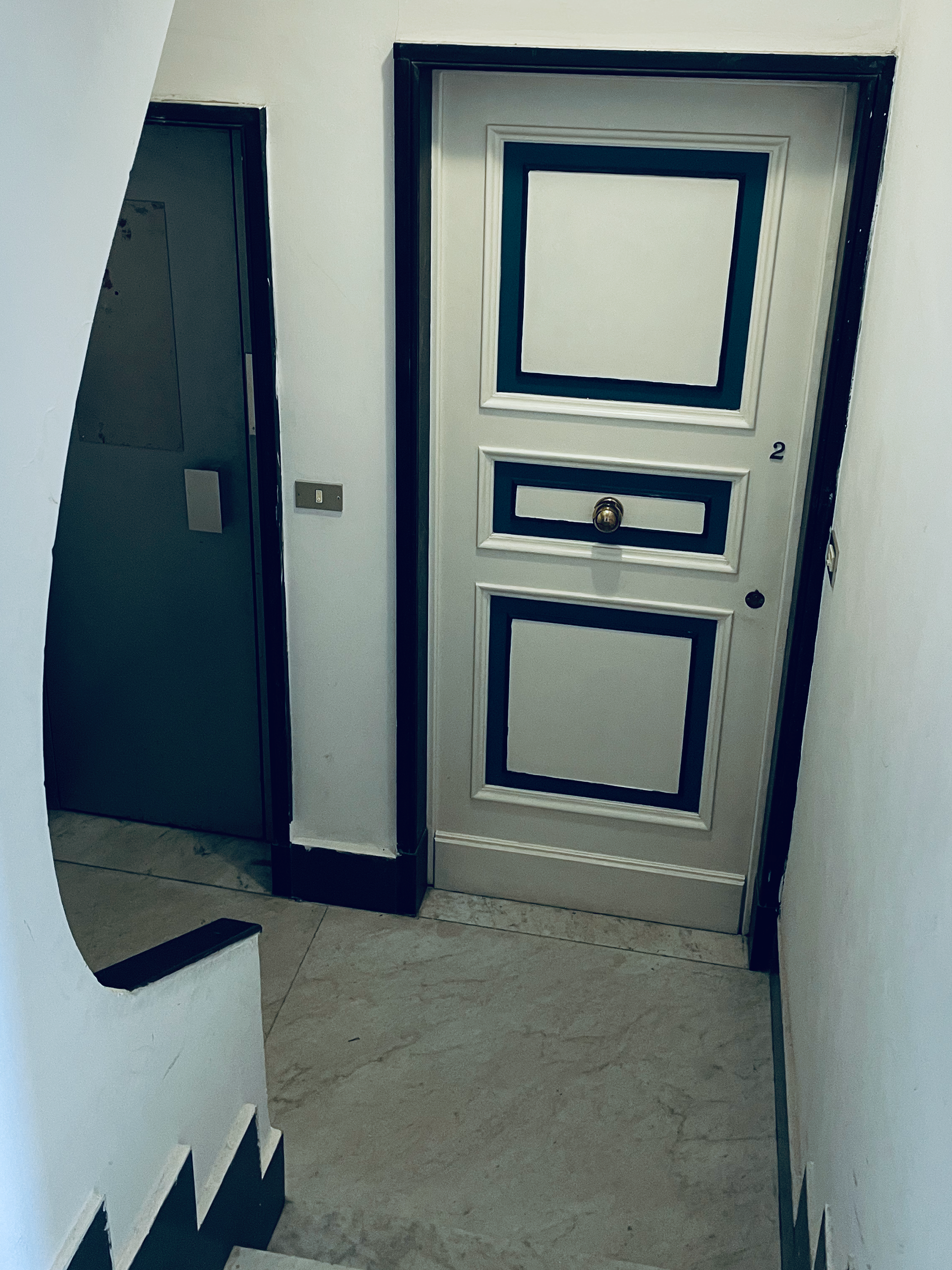 Atrio della piccola palazzina al civico 11 di Mura della Marina, nei vicoli di Genova. L'inquadratura è tagliata sulla sinistra da una tramezza dal profilo curvo. Proseguendo con lo sguardo verso destra c'è la porta metallica di un piccolo ascensore e poi il portoncino bianco dai dettagli blu notte e dal pomello dorato dell'appartamento all'interno 2. Si vede verso il basso una porzione del pavimento, perchè la foto di riferimento è scattata dall'alto di una scaletta, e un'altra del muro perpendicolare al portoncino. Il pavimento è in marmo.