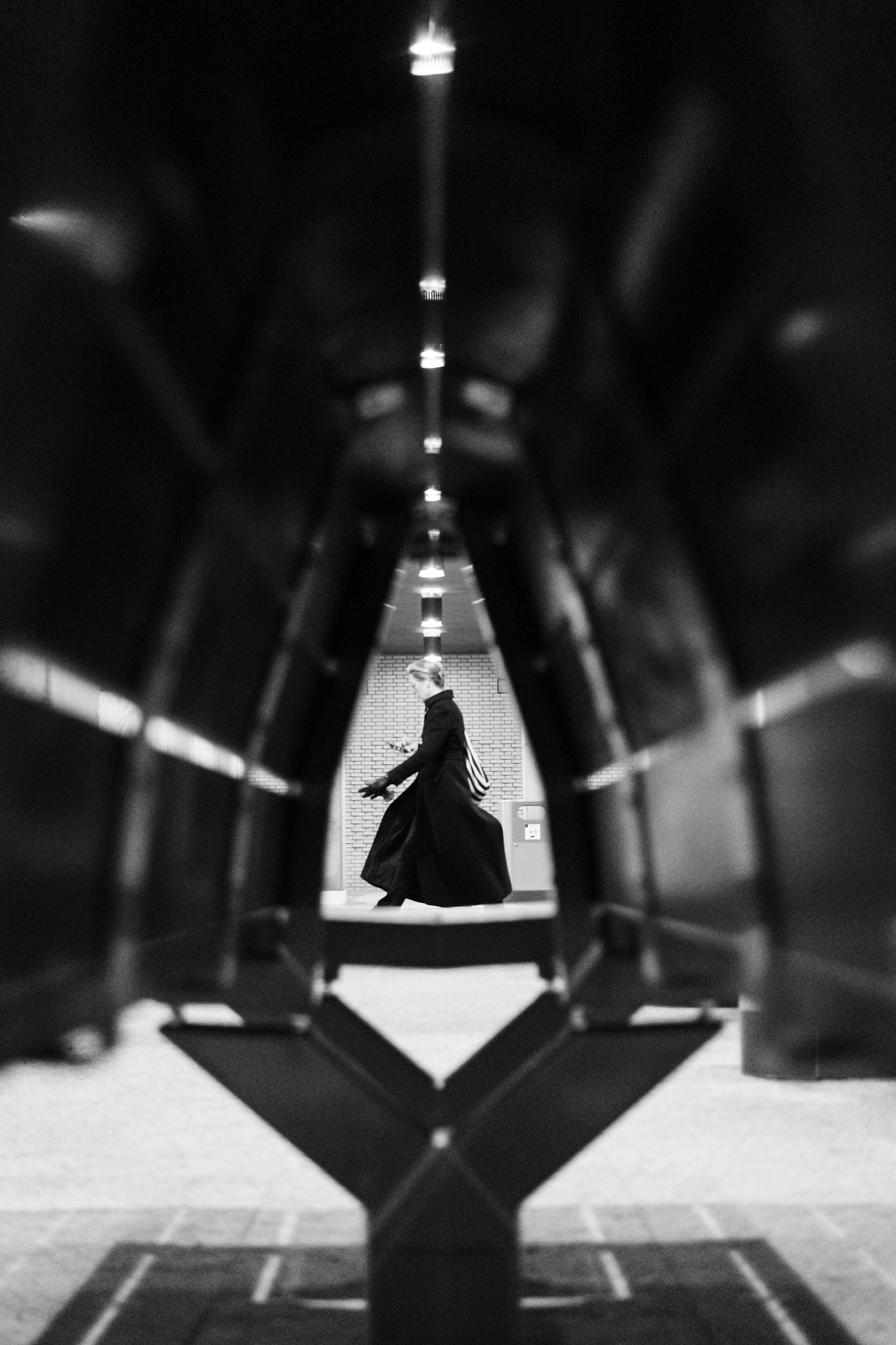 Vrouw in lange jas passeert de bankjes op Groningen Hoofdstation als Neo uit The Matrix, geframed door lichtstralen en scherpe lijnen.