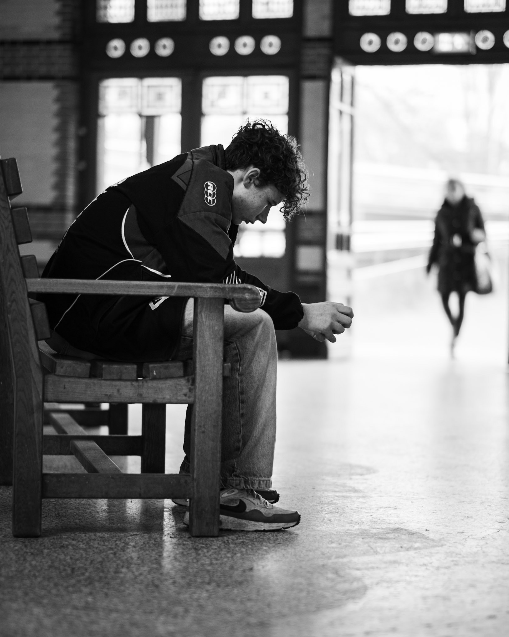 Hij zit gebogen op de houten bank, wachtend terwijl zij vervaagt in de verte.