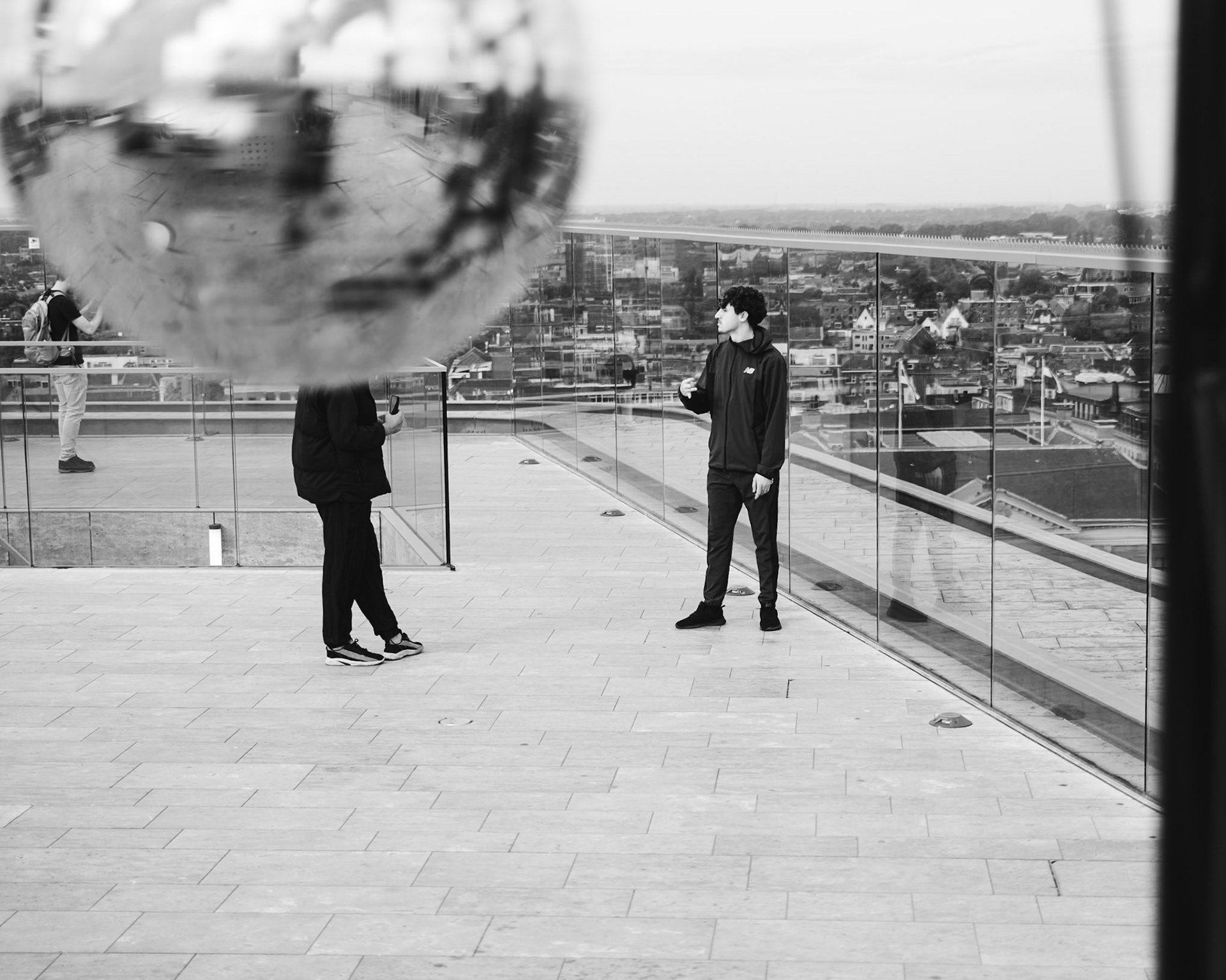 Man met discobal-hoofd converseert op Forum-dak, stad vervaagt achter glazen reling.