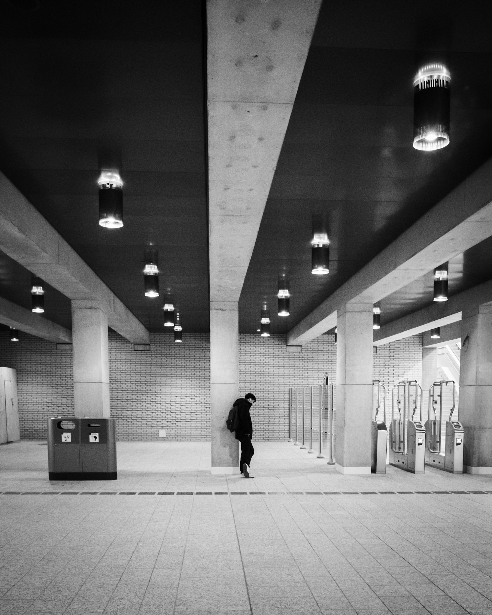 Eenzame reiziger bij de poortjes, omringd door betonnen pilaren en hangende lichten als wachters in lege hal.