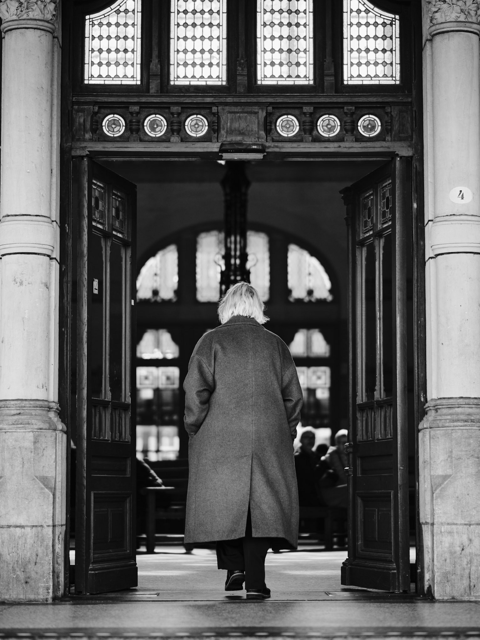 Een vrouw in lange jas stapt het historische portaal in, gevangen tussen ornamenten in een moment van vertrek.
