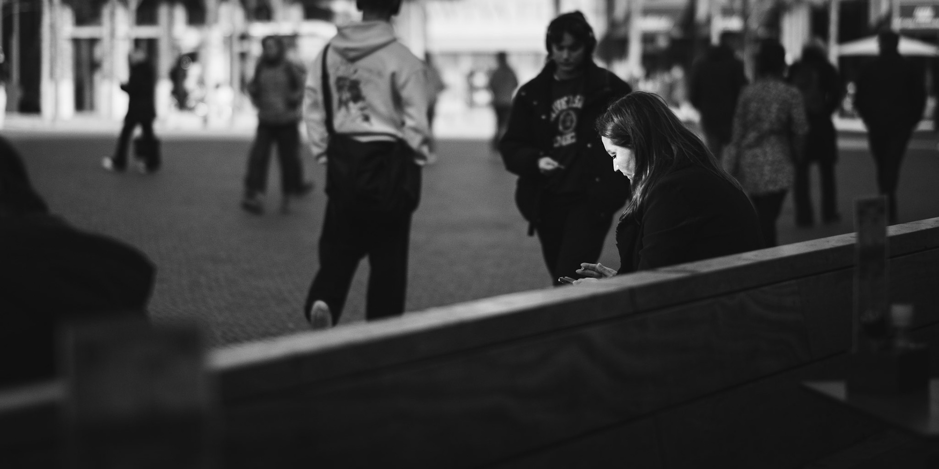 Ze zit gebogen over haar telefoon, terwijl voorbijgangers opkomen en weer vervagen in de massa.