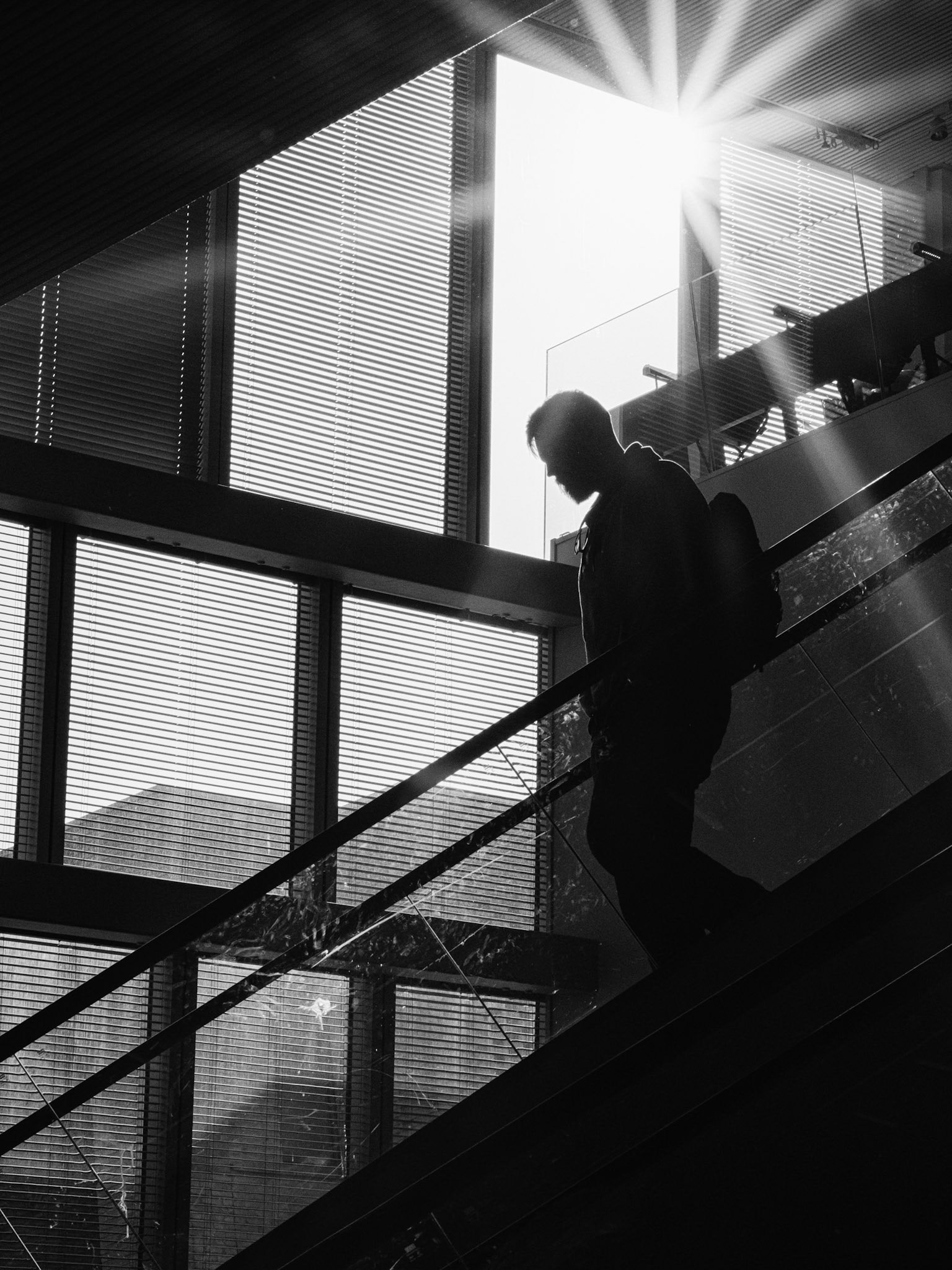 Een rugzakdrager daalt de roltrap af terwijl zonlicht door lamellen breekt, isolerend silhouet in gouden straal.