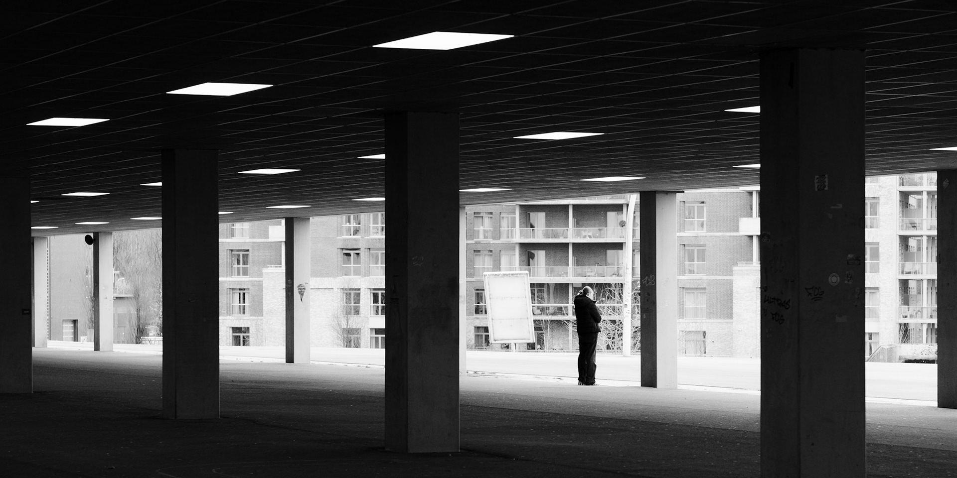 Onder Euroborg arcades: een figuur wacht, Groningen lonkt door de pilaren.