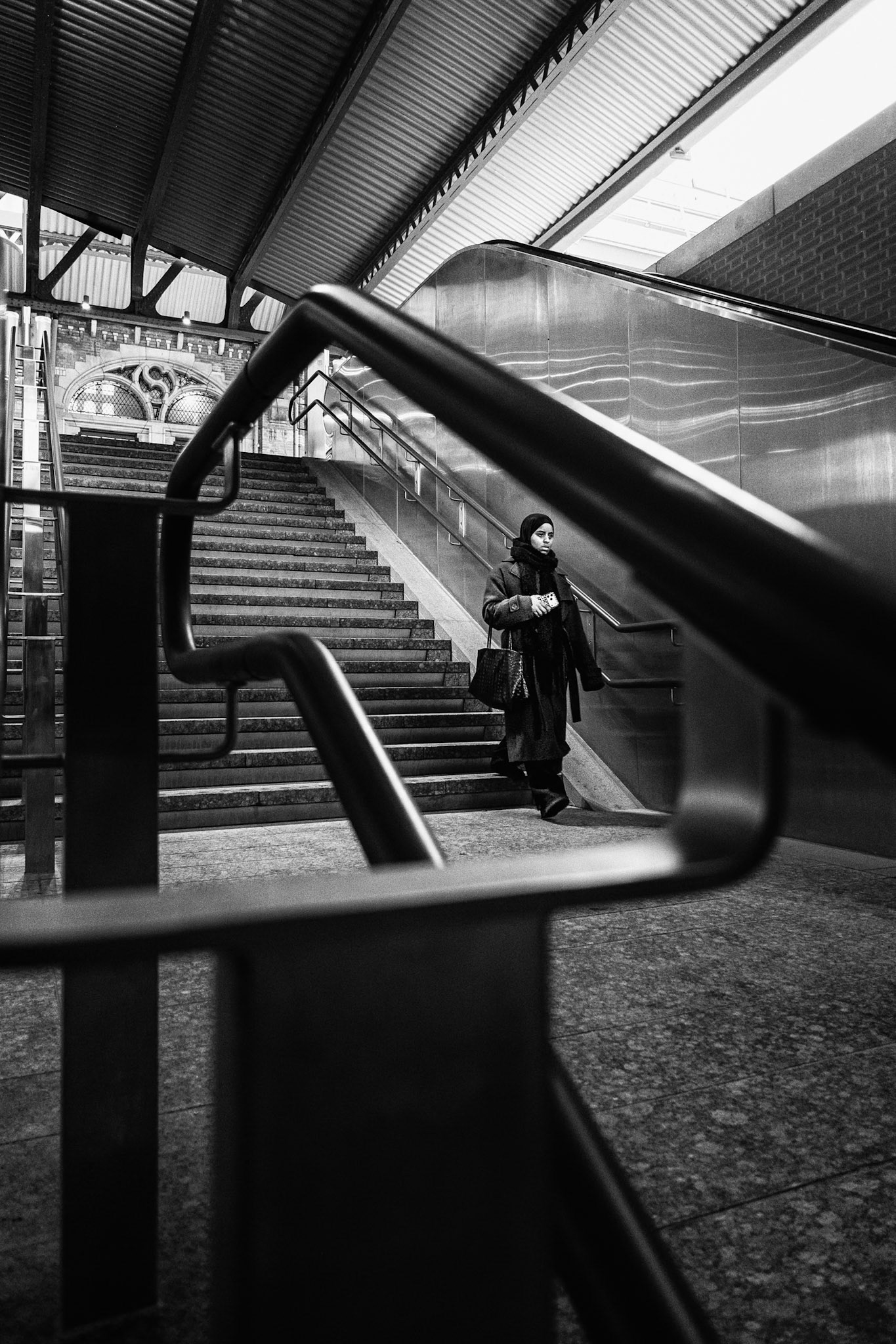 Reiziger daalt roltrap af, subframes van herhaalde leuningen en trappen vormen leading lines die figuur-ground versterken met geometrische ritme en diepte.