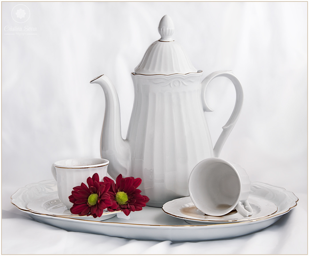 Bodegón de tetera con tazas blancas sobre fondo blanco con dos flores rojas y con una taza bolcada donde se ve el té.