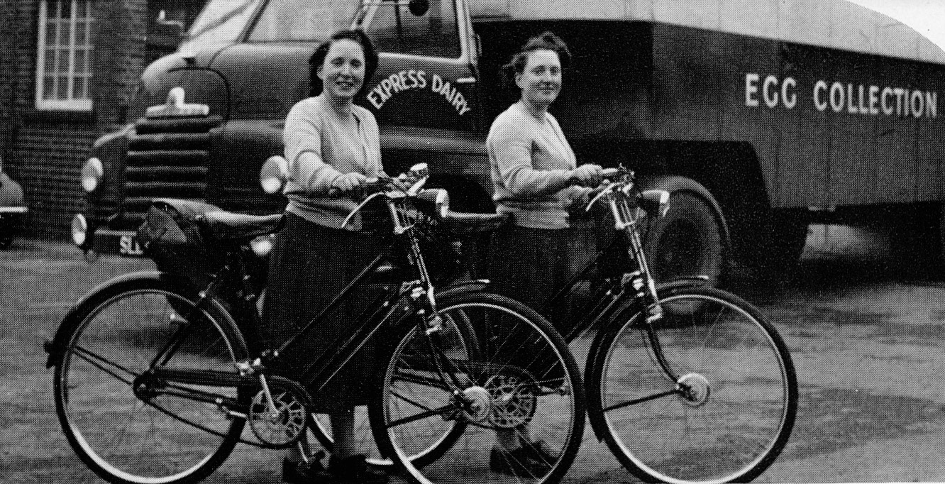1957 Peggy (left) and Doris (right) Wilkins at the Seaton Junction egg depot. Christine Richards comments "That's Peggy and Doris (Wilkins), twins who worked in Honiton Egg depot when I was there. Stayed friends till they both passed away. I was very fond of them, remember Peggy calling people who she could not remember their names Deary, I often think of them now." James Ayres comments "The vehicle in the background is an S-type Bedford tractor unit made by Vauxhall (General Motors) throughout the 1950s. Most often seen in a rigid chassis it was offered with a choice of petrol or diesel engines. The spokes of the ladies' chain-wheels spell a word. 'Triumph' perhaps?