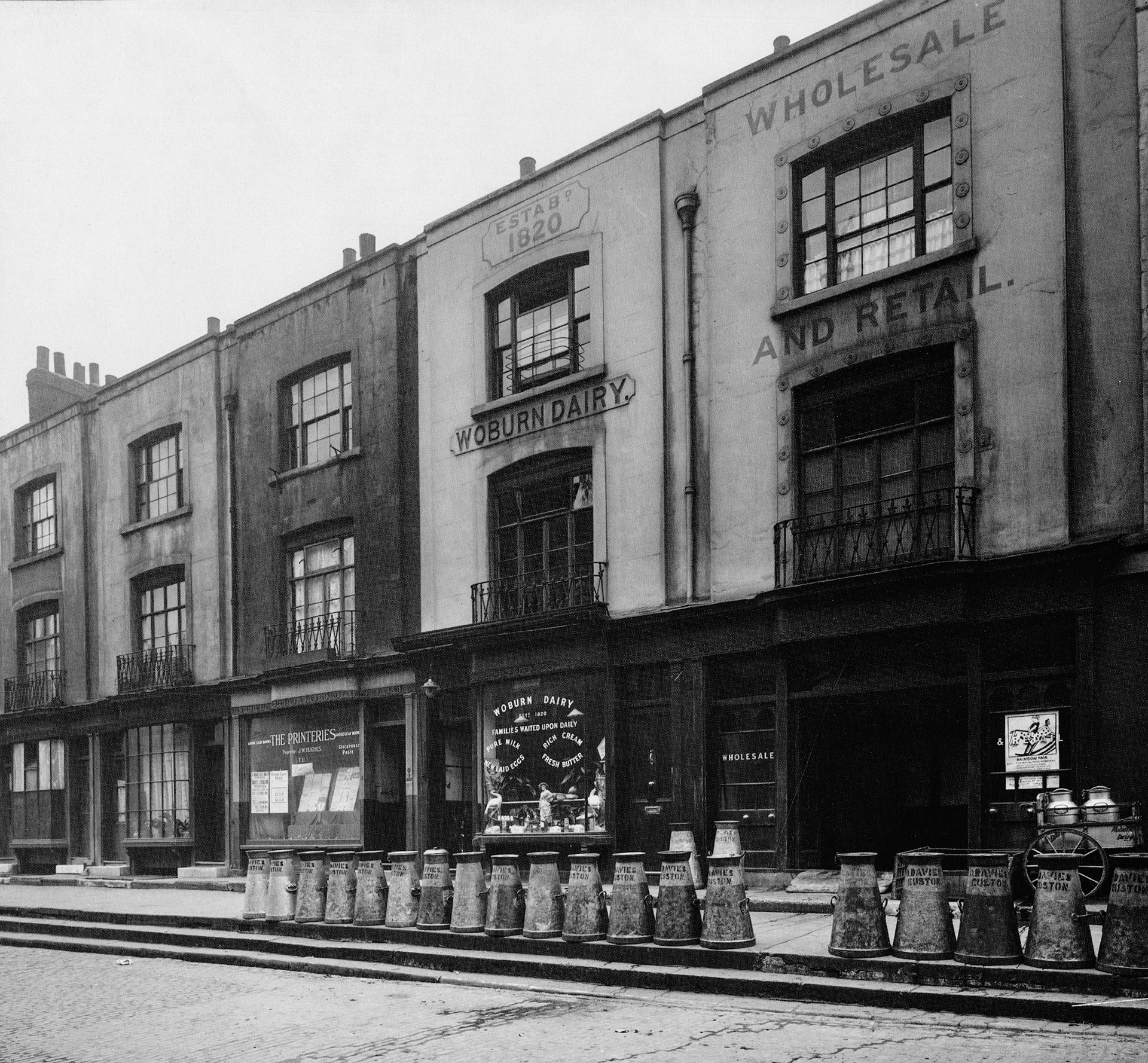 1920. Woburn Buildings, 2-14 Dukes Road, Euston, remains one of London's most picturesque places. Built to serve the grand houses on the adjacent Bedford Estate, they form part of Thomas Cubit's original design of 1822 with a beautiful sequence of curved bay windows to the shops. Milk and fresh farm produce were brought into the great railway stations daily; hence the line of milk churns on the footway, which still retains the original massive York stone slabs over the cellars beneath. (Courtesy GLC Historic Buildings Collection)