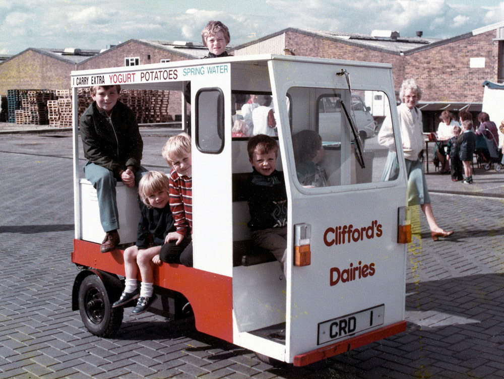 1981 This picture of kids and float was taken at an open day at Clifford’s HQ in Downshire Way, Bracknell. The float itself is tiny and there has to be a risk that the extra yoghurt, potatoes and spring water in the back caused it to do a wheelie on its rounds. The firm of Clifford’s began life in Middlesex in 1874, when patriarch William began selling milk from his two cattle door-to-door in Hounslow. This was in the days when milk was carried in pails on a yoke across the seller’s shoulders. By 1907 William had a herd of seventy cows, a farm and a number of horse-drawn milk carts. The business expanded still further between the wars and began to use dairy herds in Berkshire. Eventually the company’s offices were moved to Bracknell in 1947, and there they stayed until Unigate bought the firm in 1993 and moved operations elsewhere.