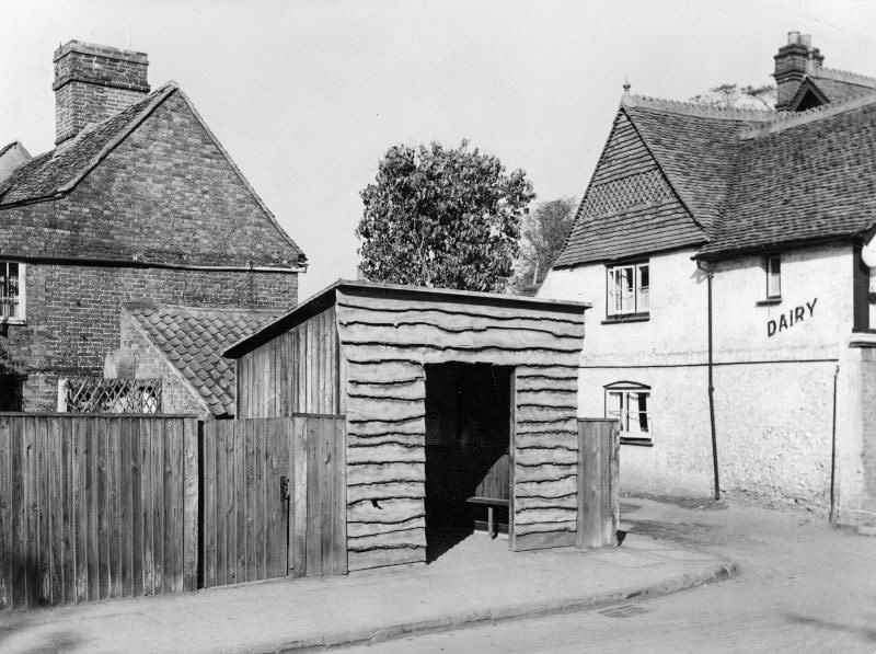 1933 Roadside bus and coach passenger shelter at Banstead, Surrey, situated on a corner alongside Hodges Dairy. David Isted comments "The corner of Salisbury Court and the old narrow High Street. It faces the plinth of the War Memorial and Park Road. The slaughterhouse is behind the dairy, situated at the eastern-most end of Banstead High Street. Beyond lies the sharp and narrow bend leading into Sutton Lane, that challenged some bus drivers, next door to the left would have been the village smithy. No evidence of these buildings survive today, most of this area was obliterated by flying bomb damage on the morning of 8th August 1944, killing an off duty soldier and injuring 32 others-altogether 28 houses and shops were severely damaged and around 200 people made homeless." (Courtesy London Transport Collection)