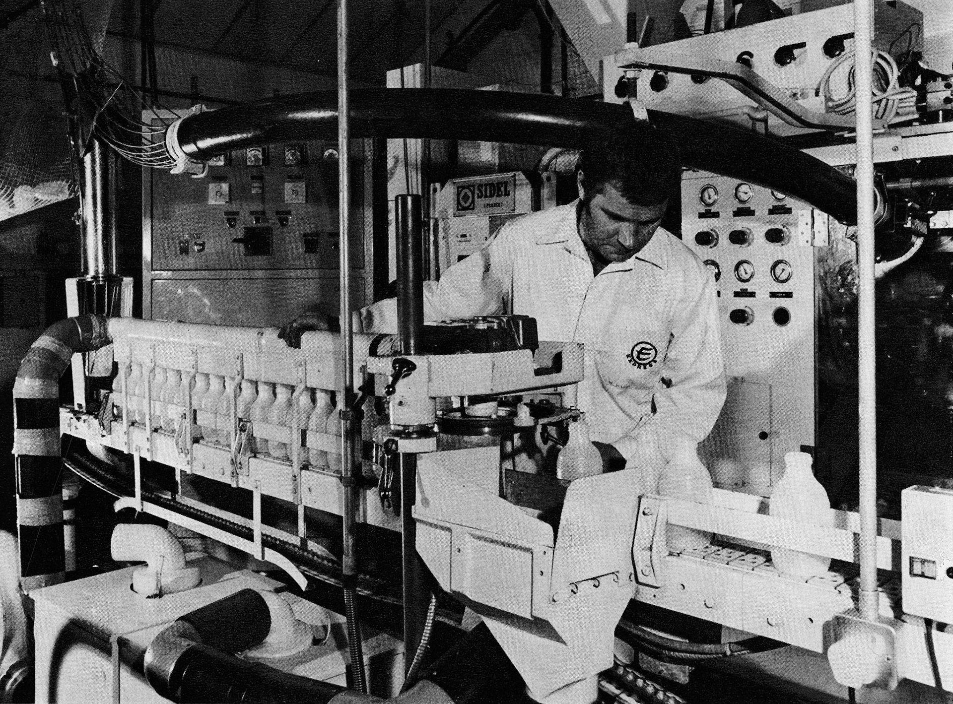 1976 Raymond Broadbent, who previously ran his own property-repairing business, watches over one of the twin French 'Sidel' blow-moulding machines. Working on a 2-shift basis, these churn out plastic 'pinties' at roughly 80 a minute, with tops trimmed, the bottles are blown several hundred yards into a huge net silo above the stacking and filling complex. (Express News Christmas)