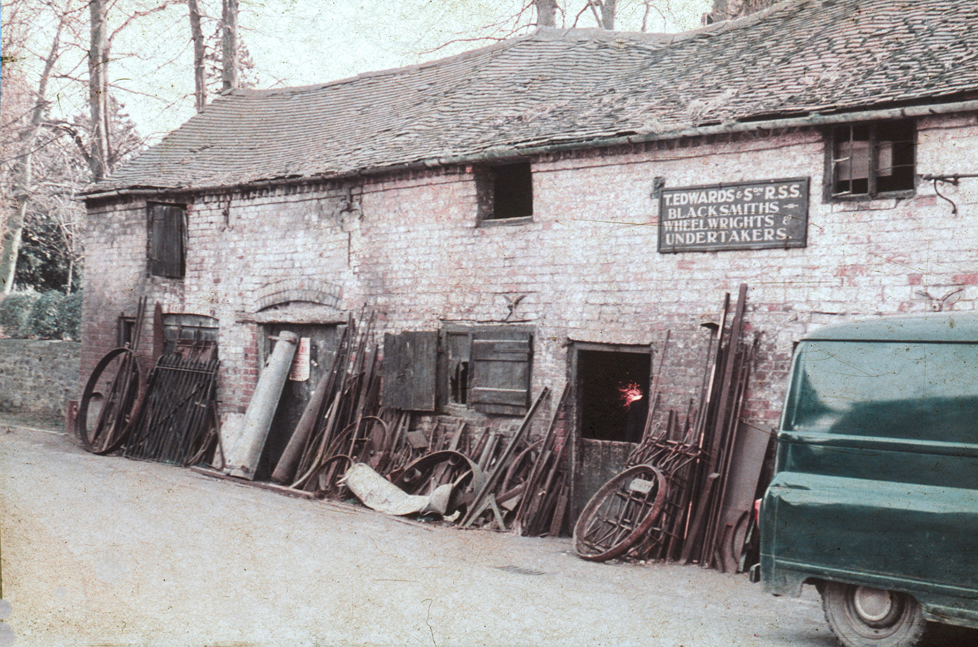 Harry Evans identified Tom Edwards, Blacksmith-he lived at Horsebridge. Jo Betts added "I remember standing at the door, watching him at the forge." Len Bailey mentions "Later, Tom Edwards had a blacksmith's shop next door-he also made coffins. As a lad I watched him shoeing a large cart horse". Chris Evans confirms that "this was opposite the Toll House; there was also a pub there till 1950's.". Mike Challinor comments "The Blacksmiths shop was on the corner of the road down to Minsterley Methodist Chapel and I would imagine it was called Horsebridge road as that is where it leads to. During my time the Blacksmith himself was Tom Edwards who actually lived at Horsebridge. If my memory serves me right he was also an undertaker. I know he had at least one son, Glyn Edwards, who was a little older than me but he was at Priory Boys the same time as me." Yvonne Davies adds "Mr Bowens' green van from the shop. (Joe Lyons 35mm slides)